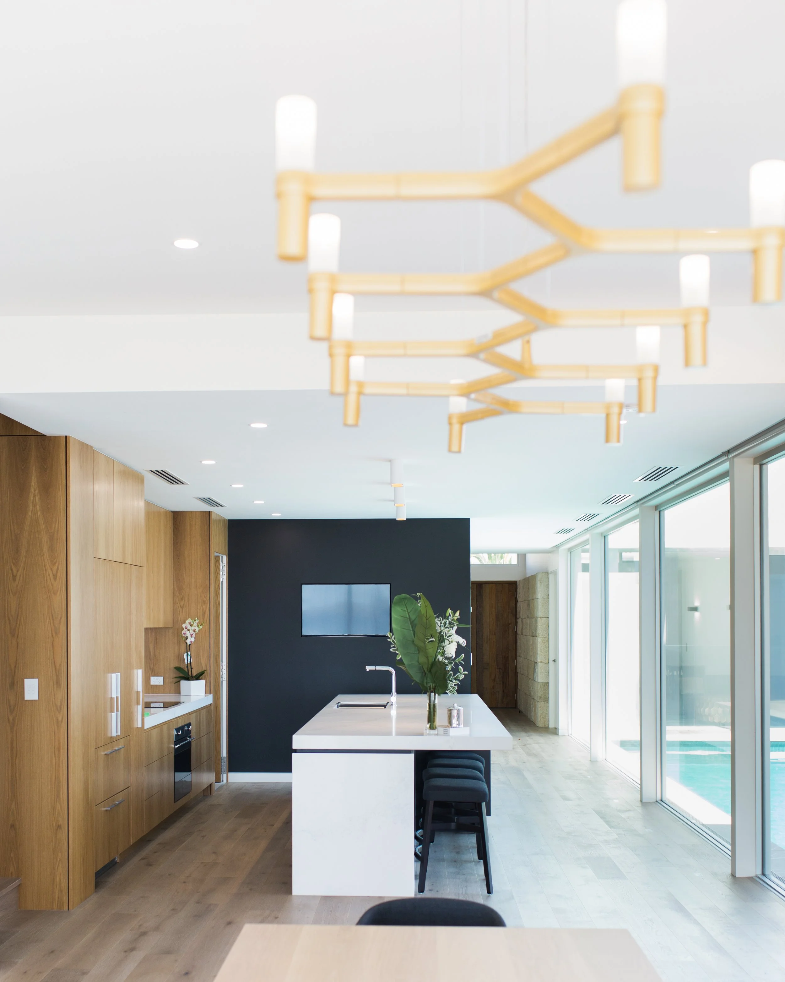 Modern kitchen with a white island, wooden cabinets, black wall, large window with sliding doors, and minimalist chandelier.