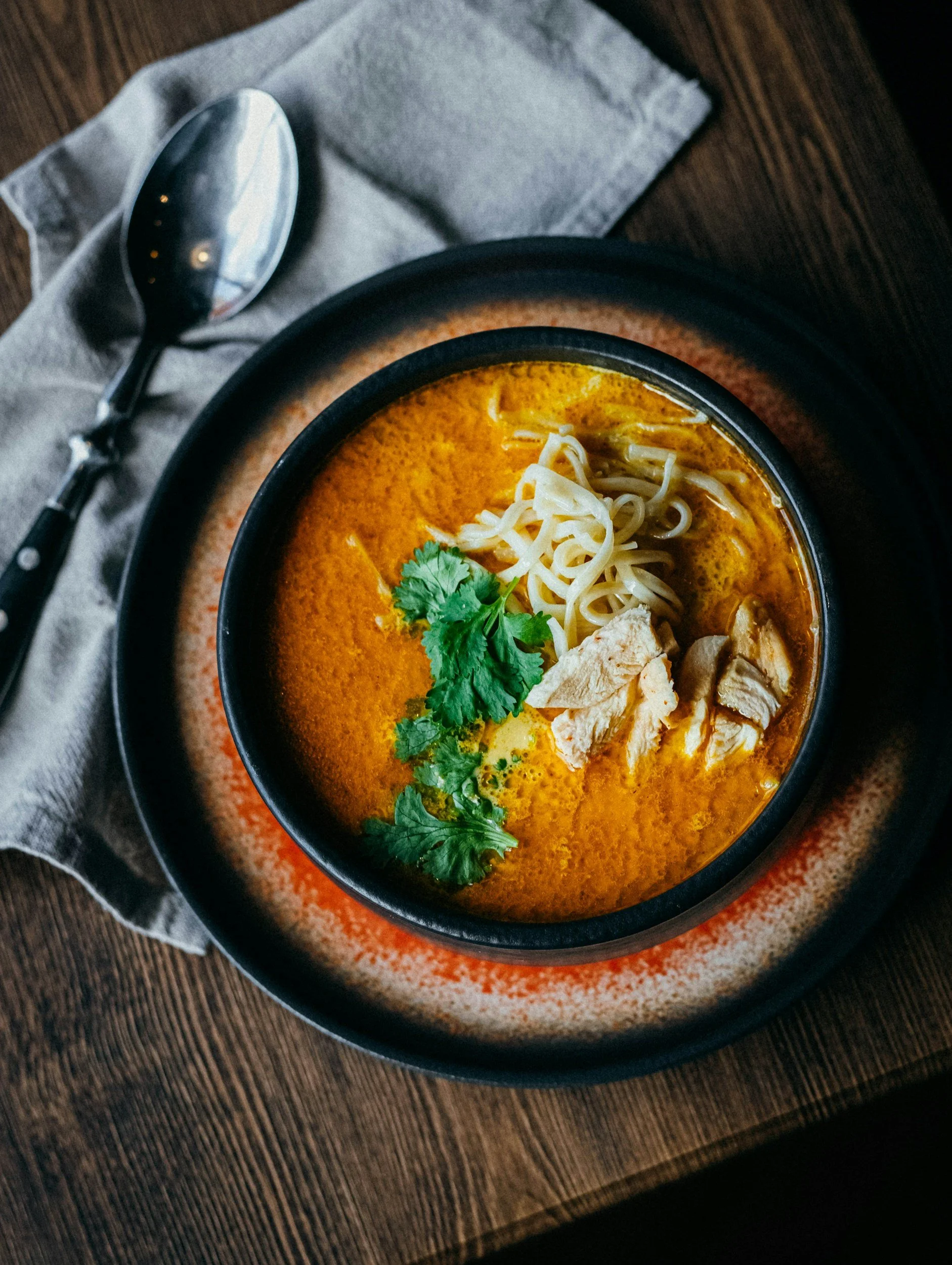 A bowl of spicy Thai curry soup garnished with cilantro, shredded chicken, and noodles, served on a black plate with a spoon and a napkin on a wooden table.