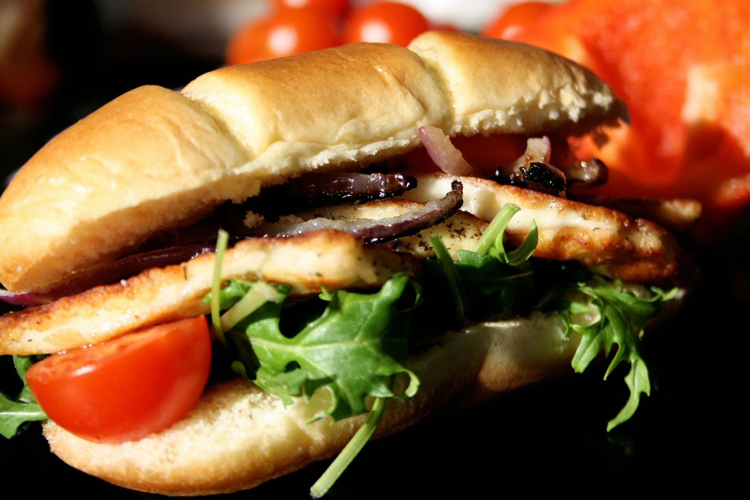 Close-up of a sandwich with grilled chicken, lettuce, tomato, onions, and melted cheese in a bun, with tomatoes in the background.