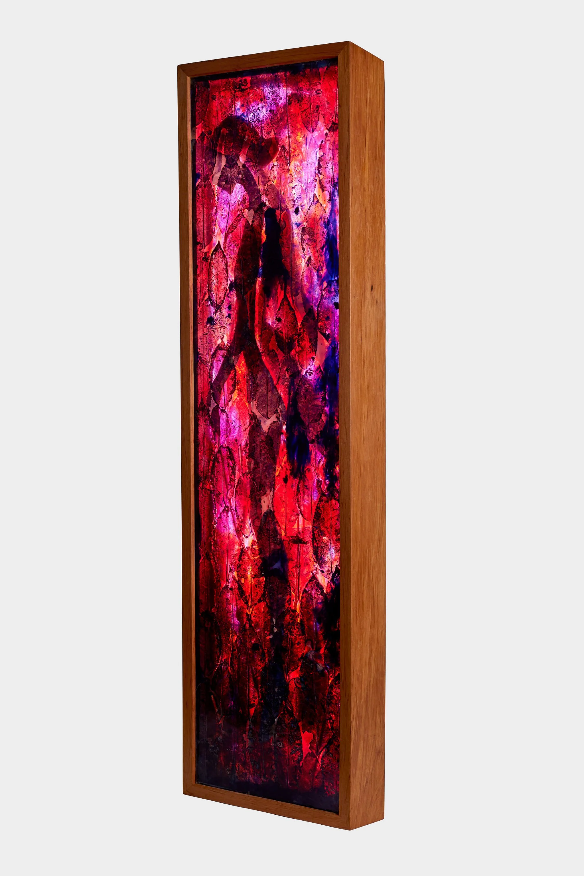 A tall vertical framed artwork with mahoe leaves in resin and a woman figure in shades of red, pink, purple, and black