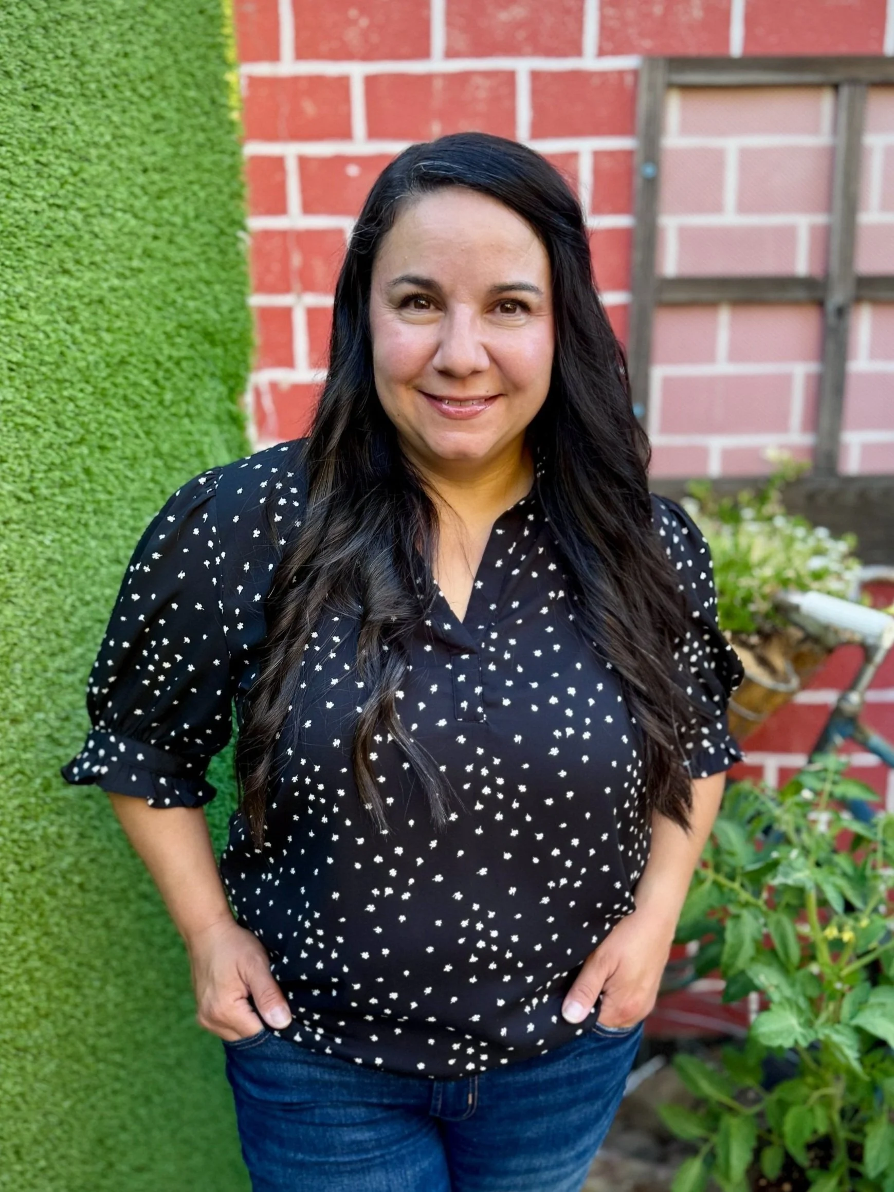 A woman with long dark hair, wearing a black blouse with white polka dots and blue jeans, smiling while standing outdoors near a green hedge and brick wall.
