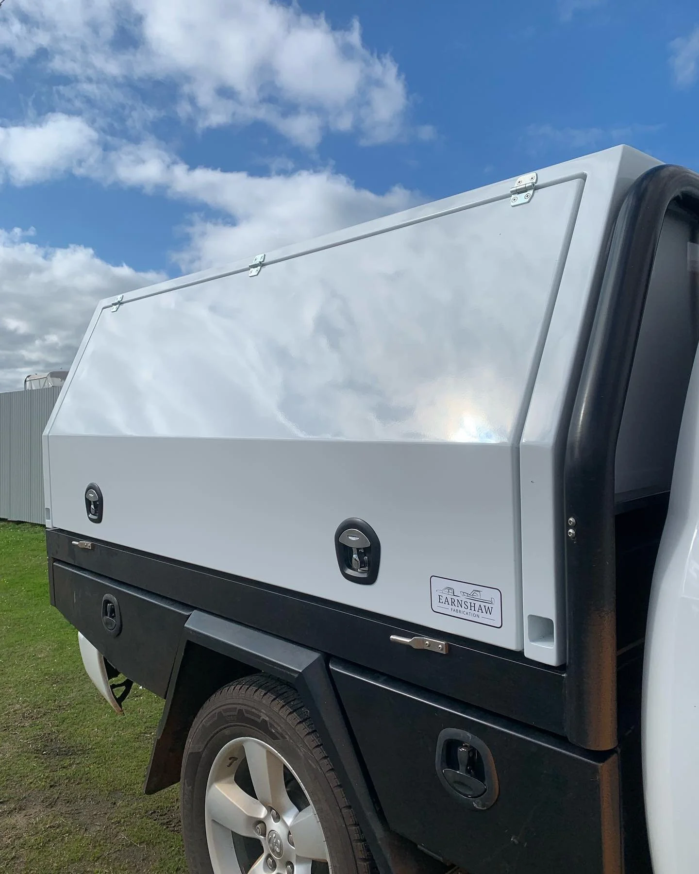 A white truck with a secure, lockable storage compartment on the side, parked outdoors under a partly cloudy sky.