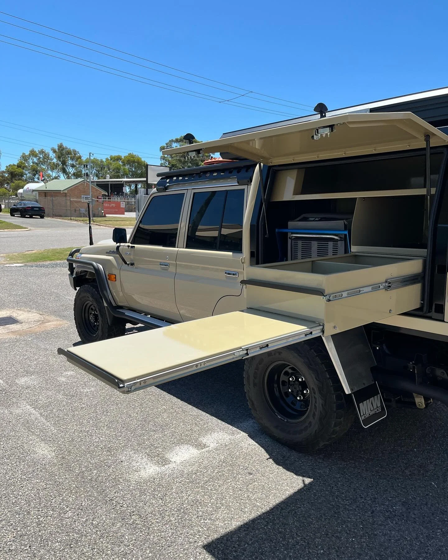 A modified beige four-wheel drive vehicle with an extended cargo area and a pull-out tray, parked outdoors on a sunny day with clear blue skies.