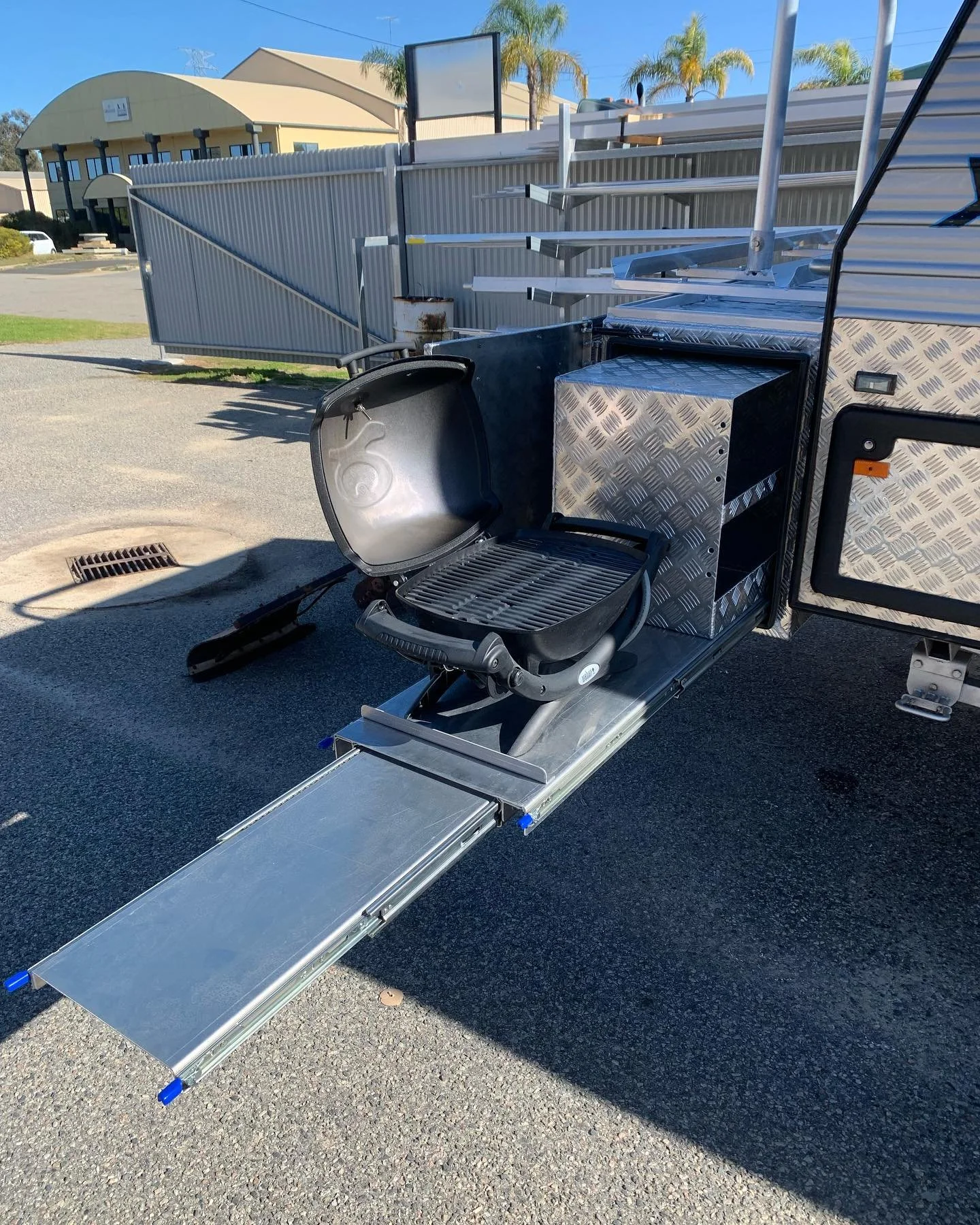 A mobile food truck with a metal ramp extended, a black folding chair inside, and a silver storage cabinet. There are metal shelves on the side and a basketball hoop in the background.