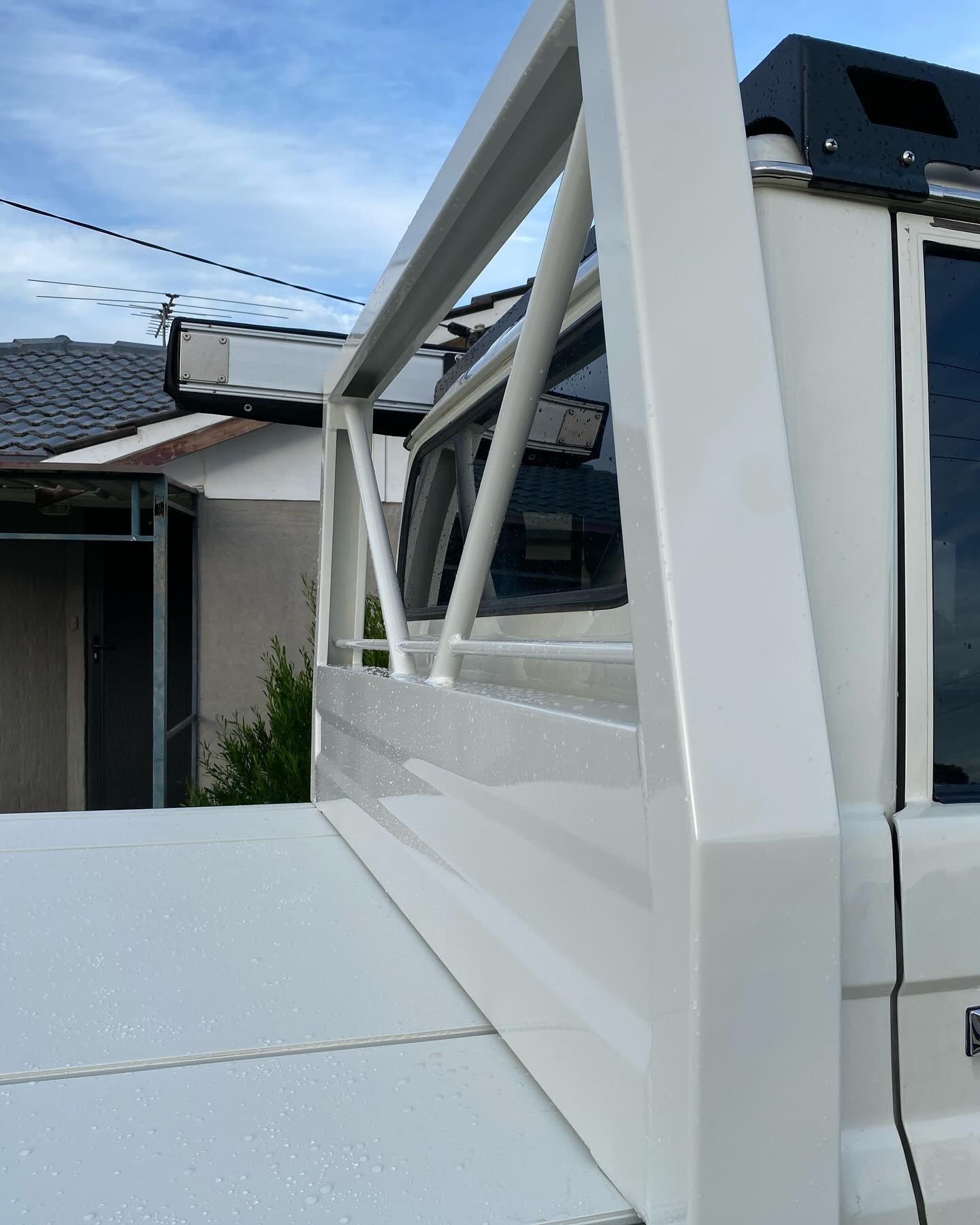 Close-up of a white utility truck with a metal frame attached to the side, water droplets on the surface, in front of residential houses under a partly cloudy sky.
