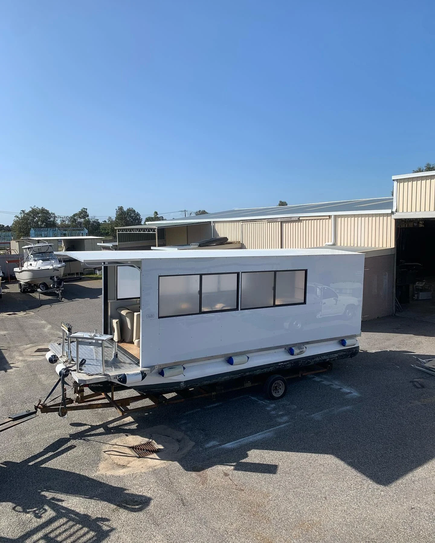 A white mobile food or coffee trailer parked outdoors near a warehouse, with windows, an open side serving area, and a small seating area inside.
