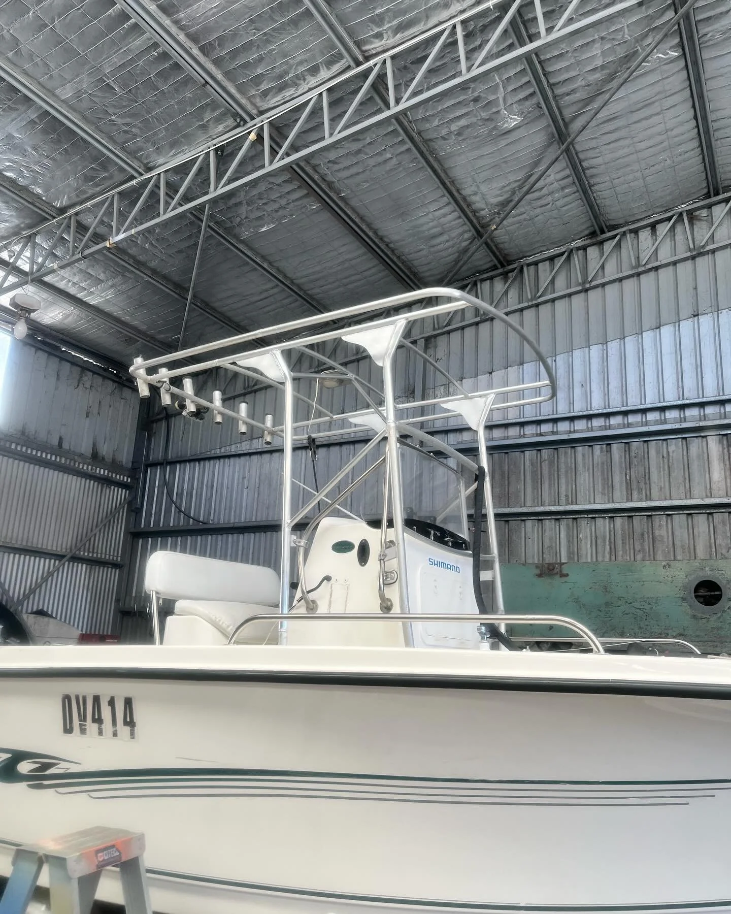 A white fishing boat with a T-top structure, parked inside a metal storage building, with an outboard motor labeled 'Shimano' at the stern.