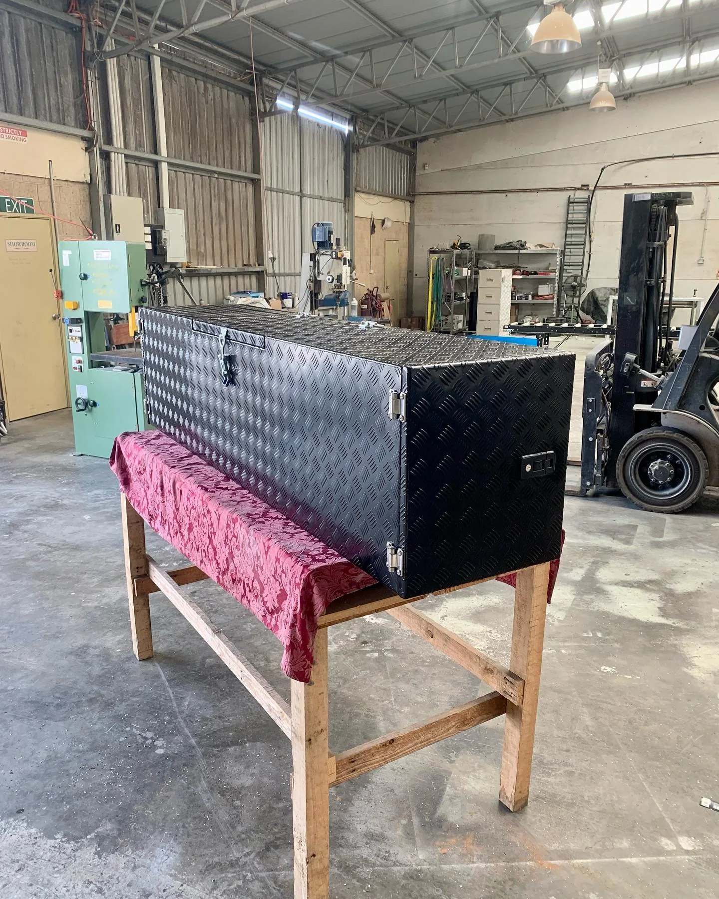 A large black diamond-plate tool storage box on a wooden workbench covered with red fabric inside a workshop or industrial garage with various tools and machinery in the background.