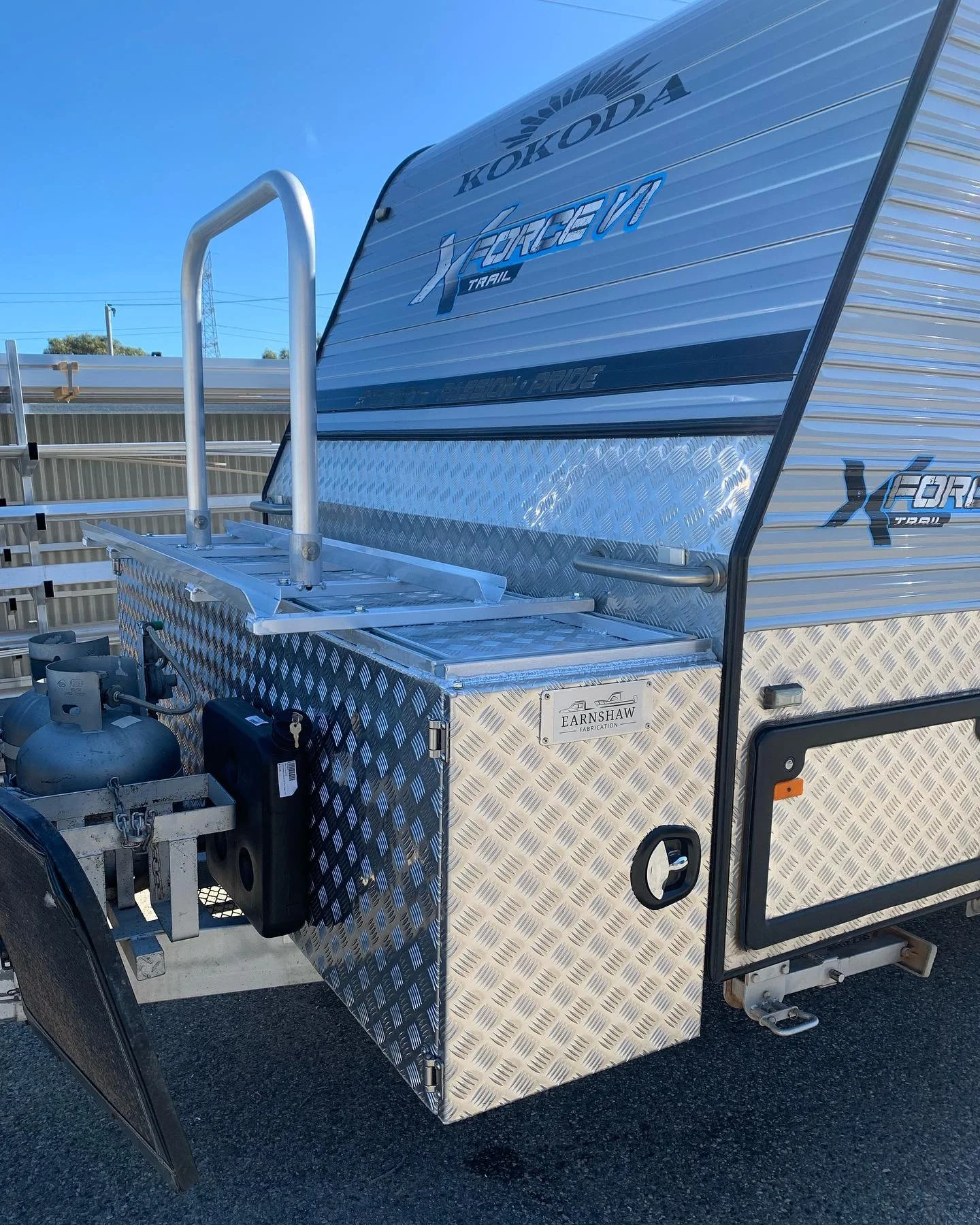 A metal travel trailer with branding that reads 'Kokoda' and 'X-Force' is parked outdoors. It features a diamond plate storage box, a propane tank, a lockable panel, and a step with a handle, all set against a clear blue sky.