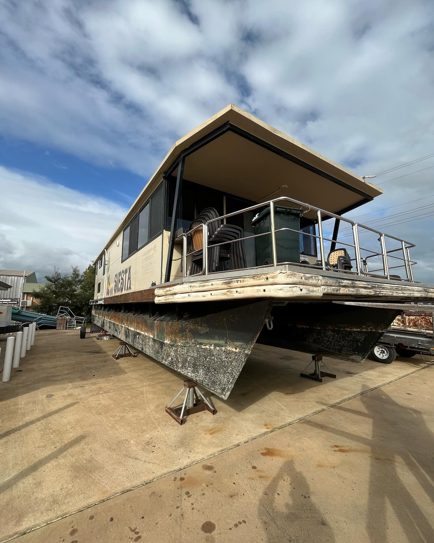 A houseboat on dry land with visible rust and algae on its hull, supported by metal stands, with a clear sky and some buildings in the background.