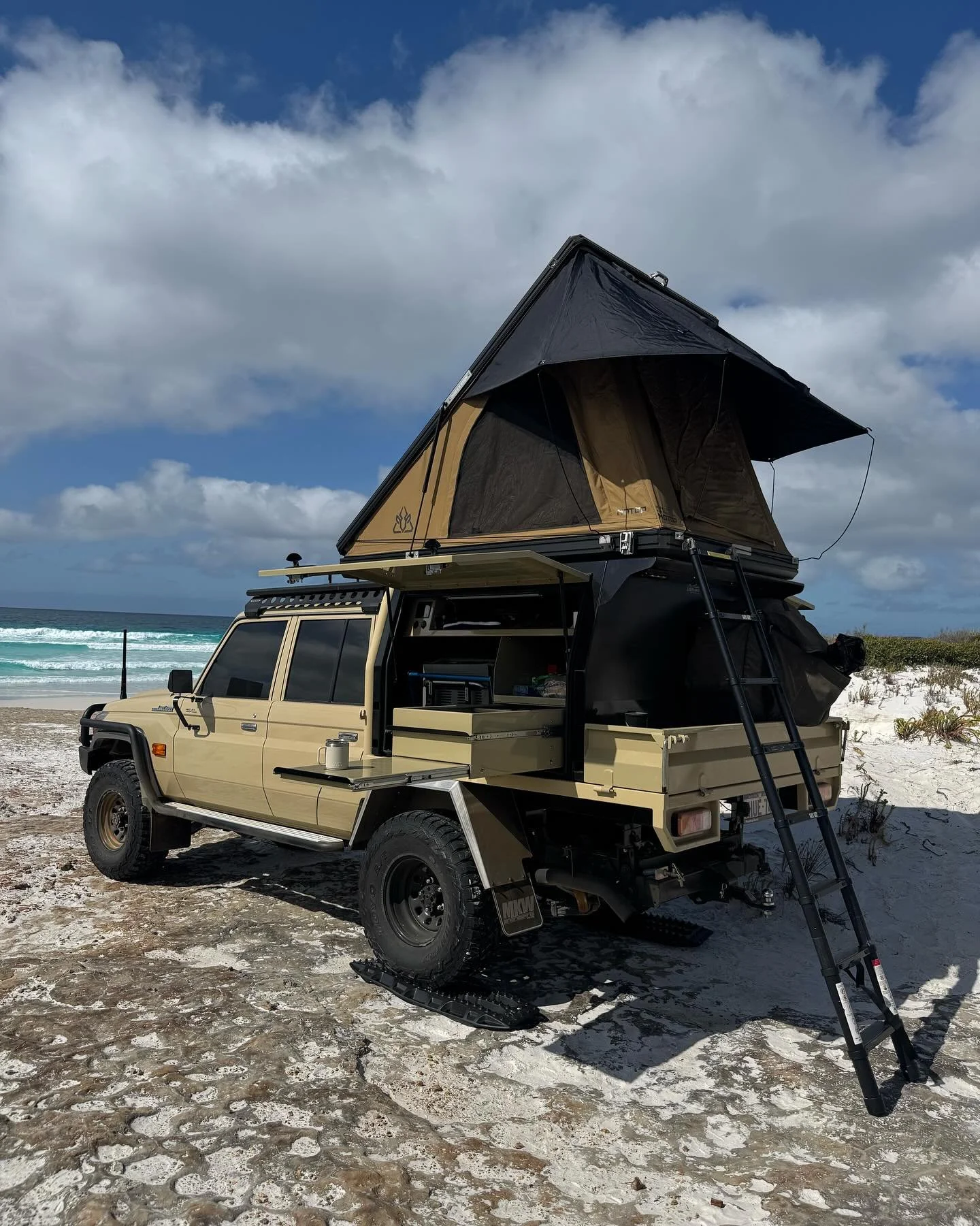 Off-road vehicle with rooftop tent set up on a sandy beach with ocean and sky in the background.