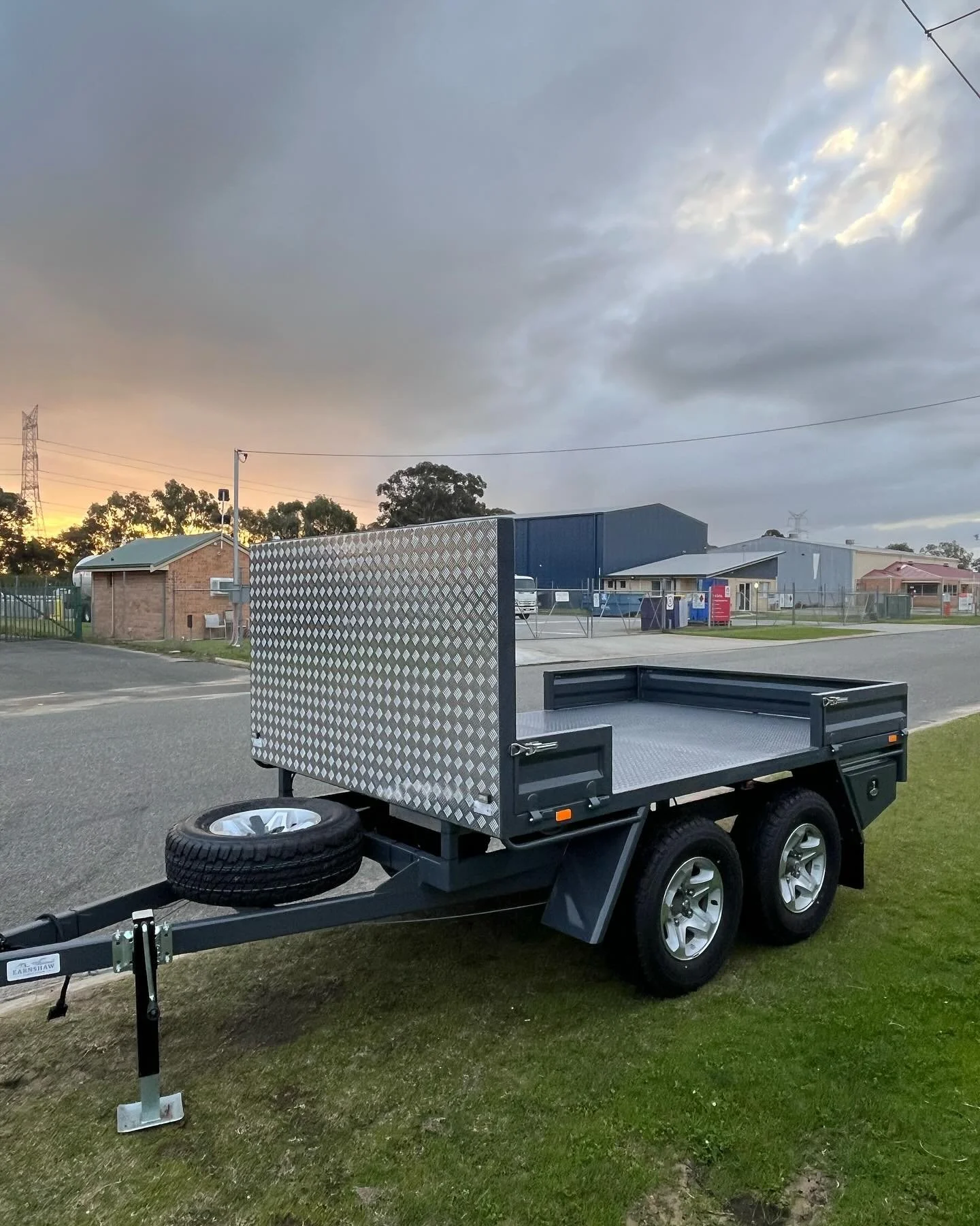 A utility trailer with black sides, silver wheels, and a diamond plate back gate, parked on grass at sunset on a street with industrial and residential buildings in the background.