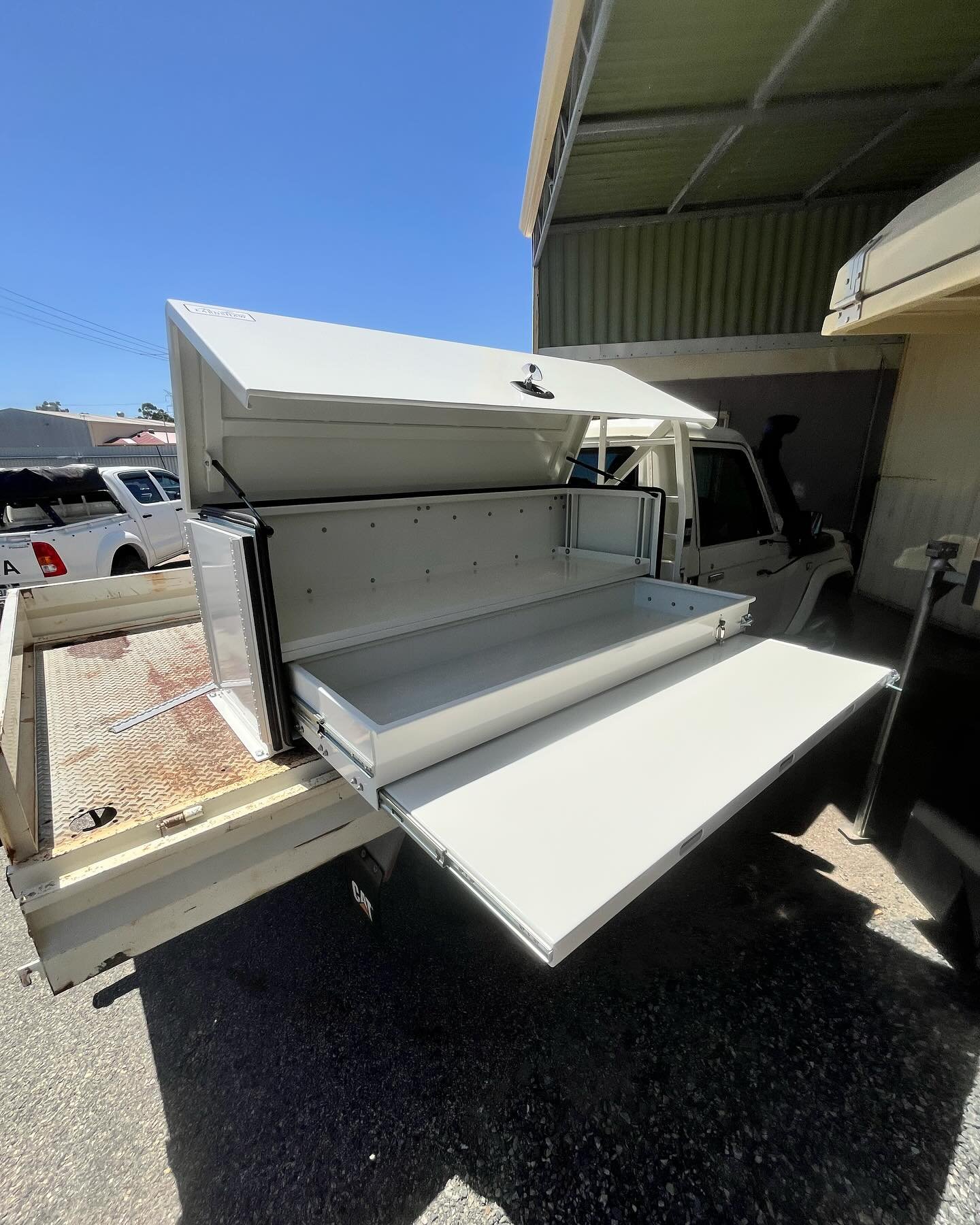 White utility truck with open storage compartments and fold-down work surface, parked under a metal shelter on a sunny day.