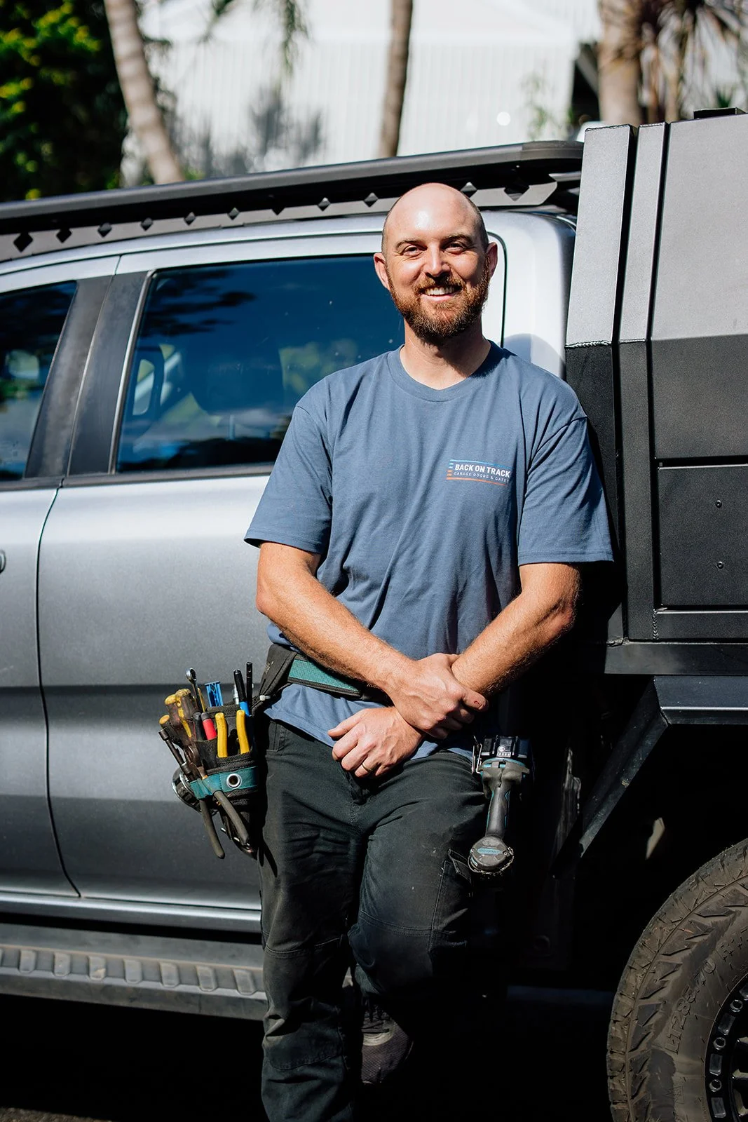 Ash from Back On Track Garage Doors is a smiling man with a beard and a bald head. He leans against a silver truck in an outdoor setting. He is wearing a blue t-shirt with the Back On Track logo, black pants, and has a tool belt with various tools.
