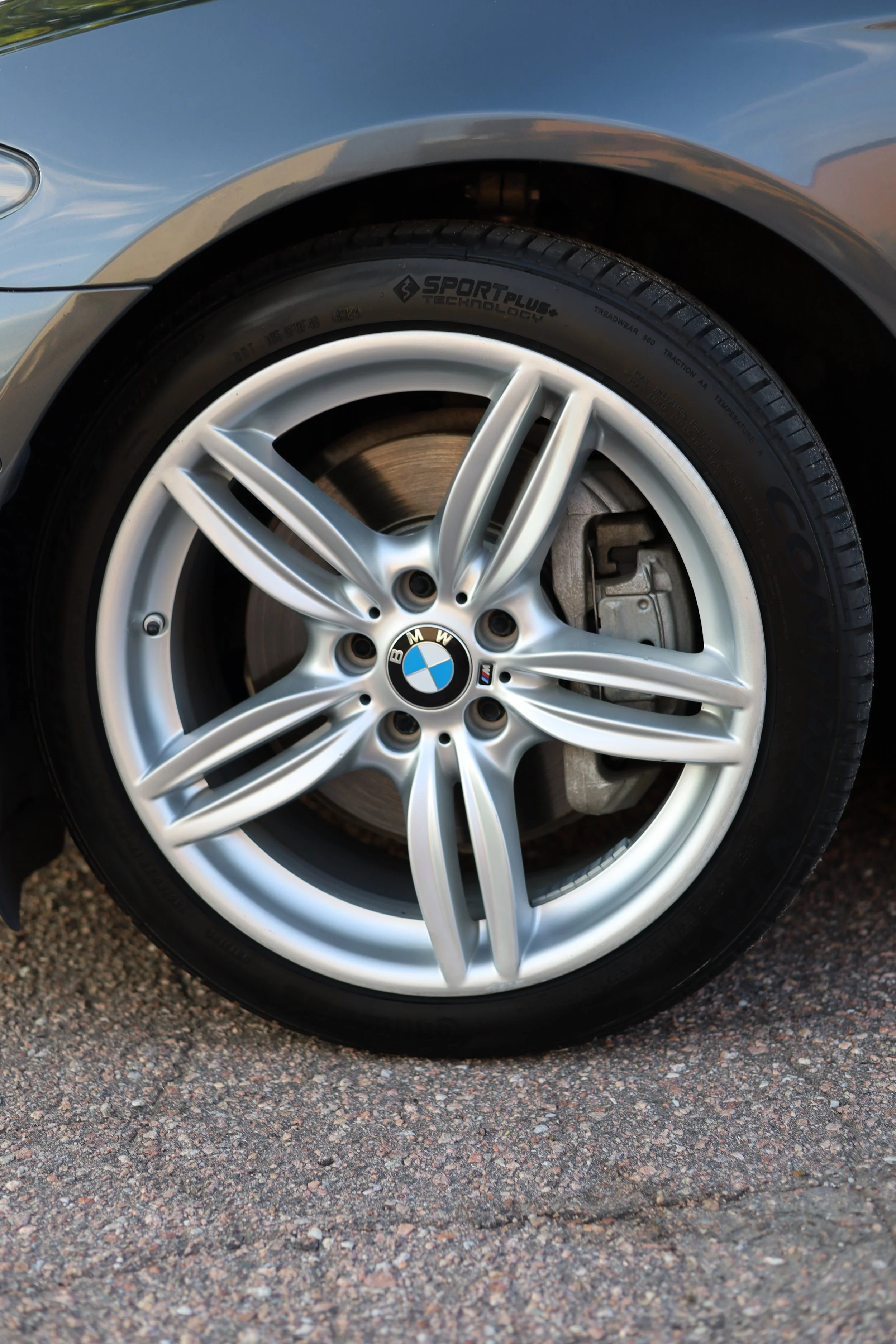 Close-up of a BMW car wheel with a silver alloy rim and black tire, showing the BMW logo at center, on a gravel surface.