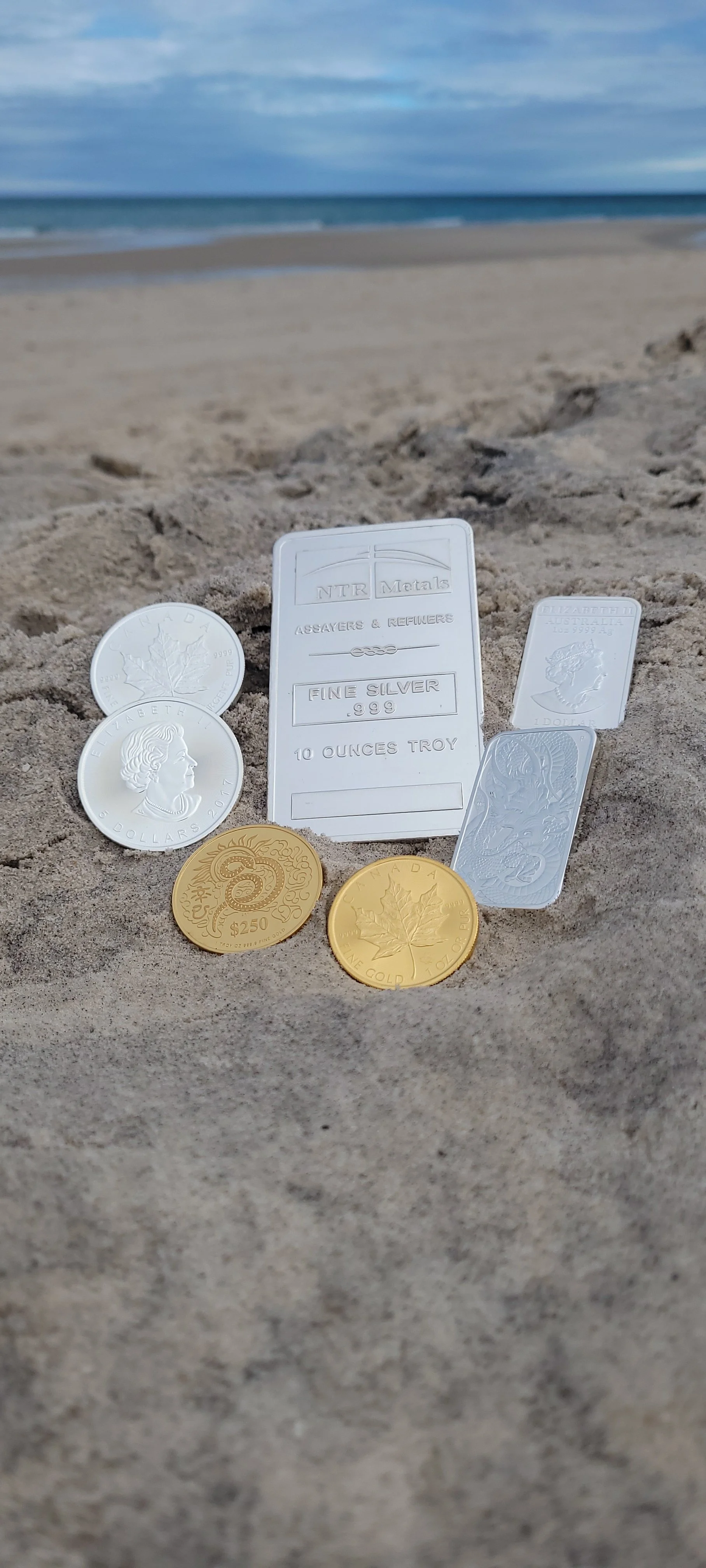 Collection of silver and gold coins, a silver bar, and silver bars on sandy beach with ocean and cloudy sky in background.