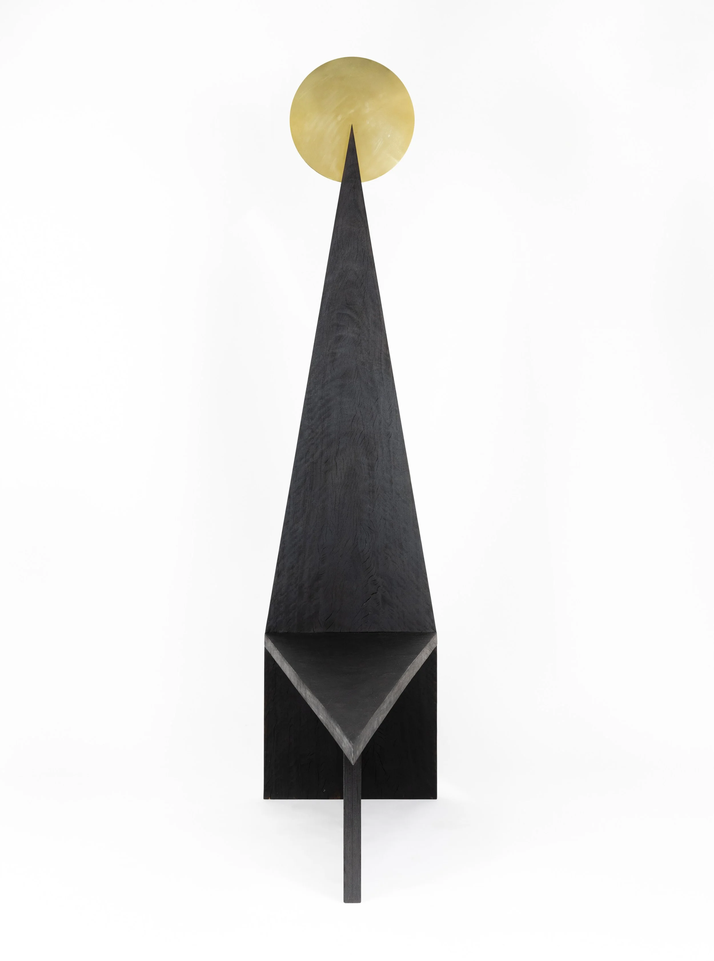 A modern, black geometric sculpture with a triangular shape at the top and a circular golden element above it, against a plain white background.
