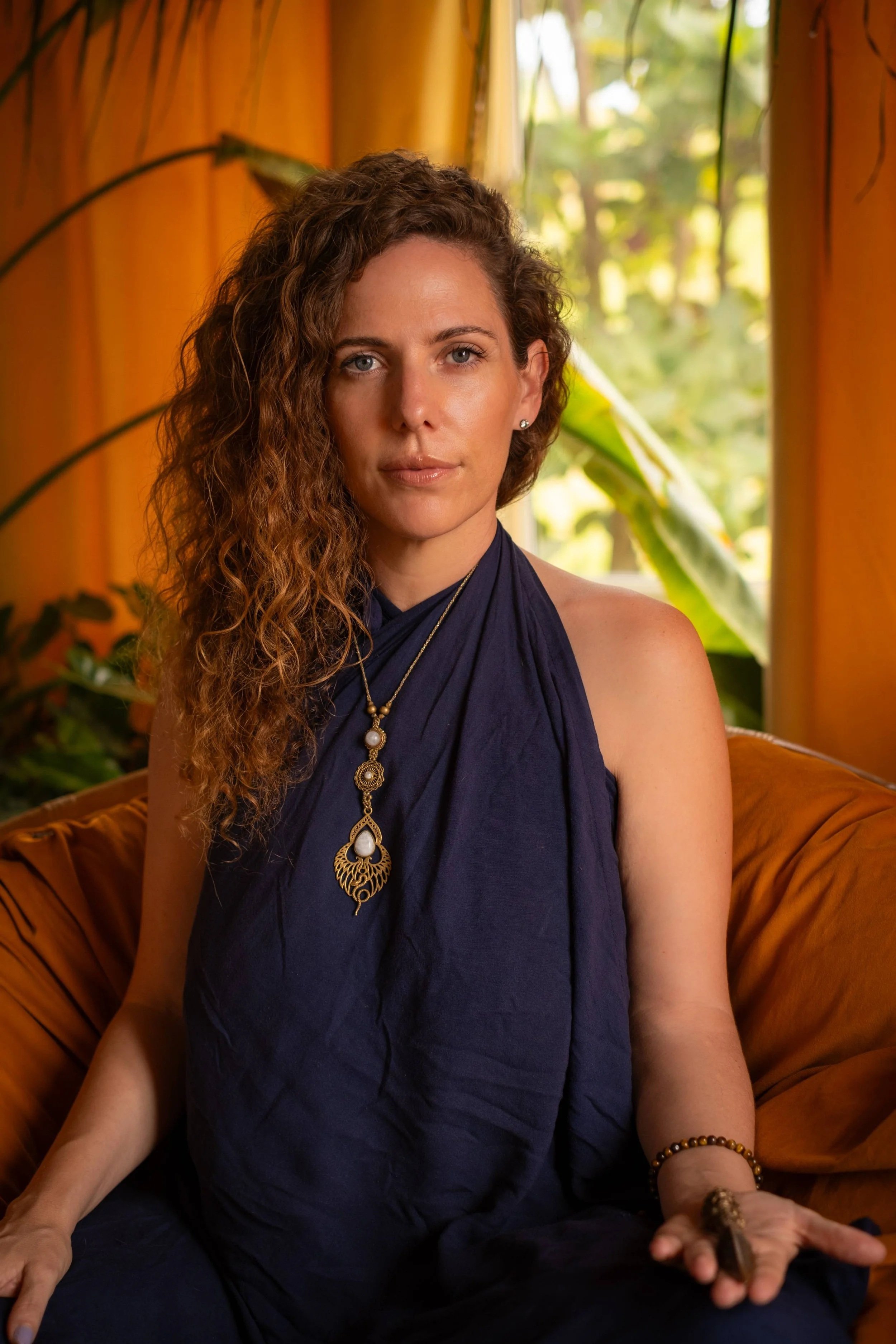 A woman with curly brown hair and blue eyes sitting on an orange couch in front of a window with green plants outside. She is wearing a dark blue sleeveless top, gold jewelry, and bracelets. She holds an object in her right hand and has a relaxed expression.