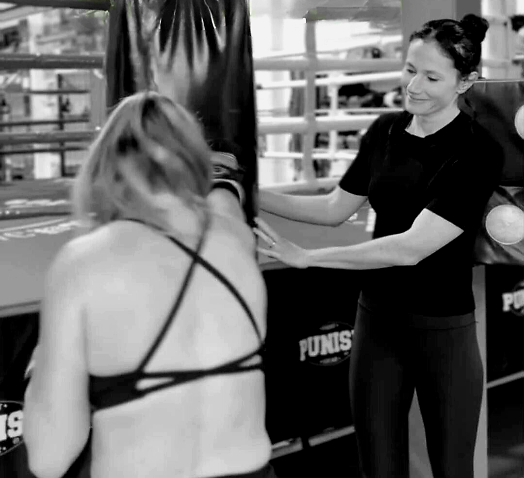Boxing gym with two women, one coaching and one in workout gear, near boxing equipment.