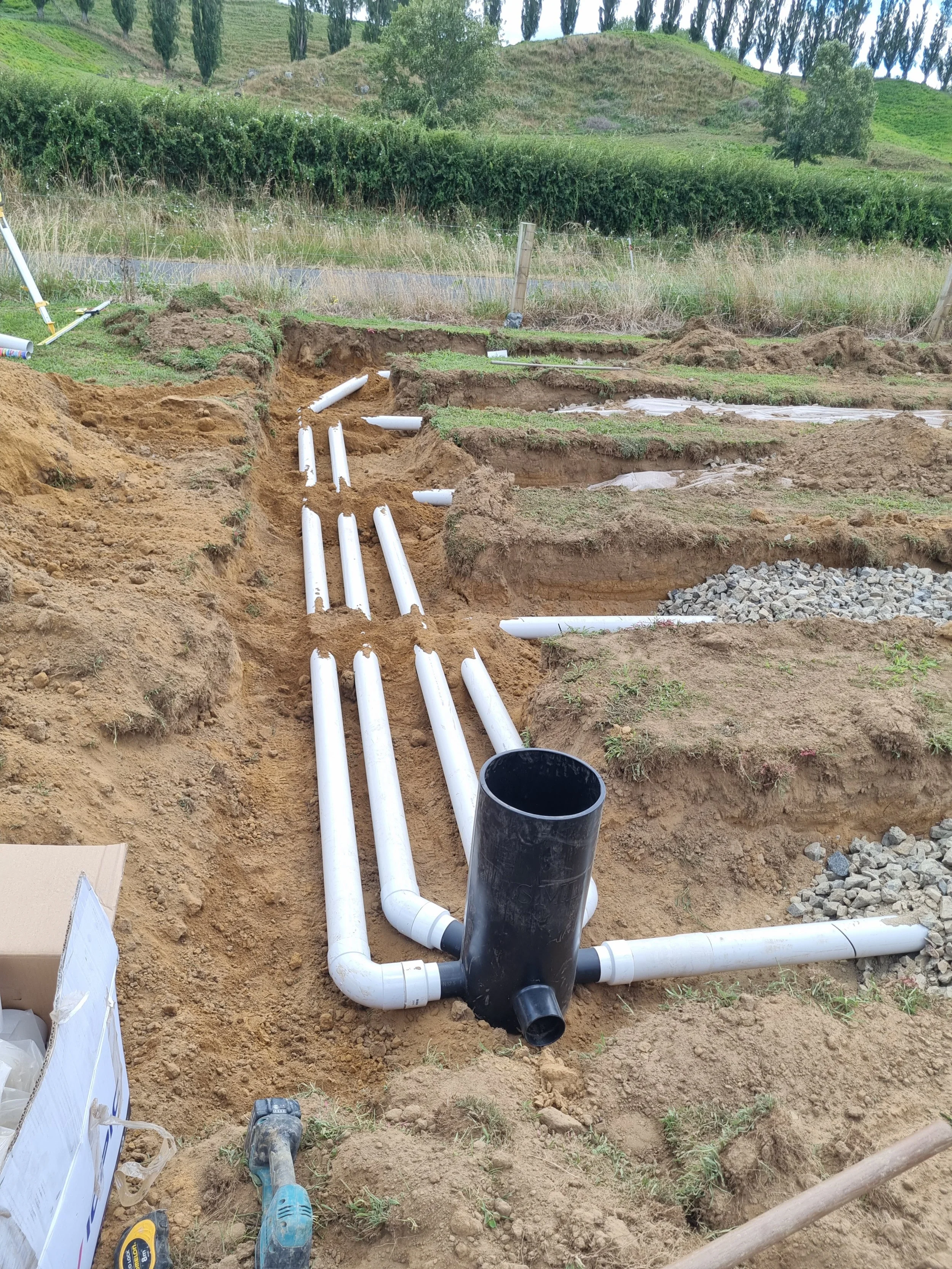 Underground plumbing installation with white PVC pipes and a black sump pump in a dirt trench at a construction site, with grassy landscape and trees in the background.