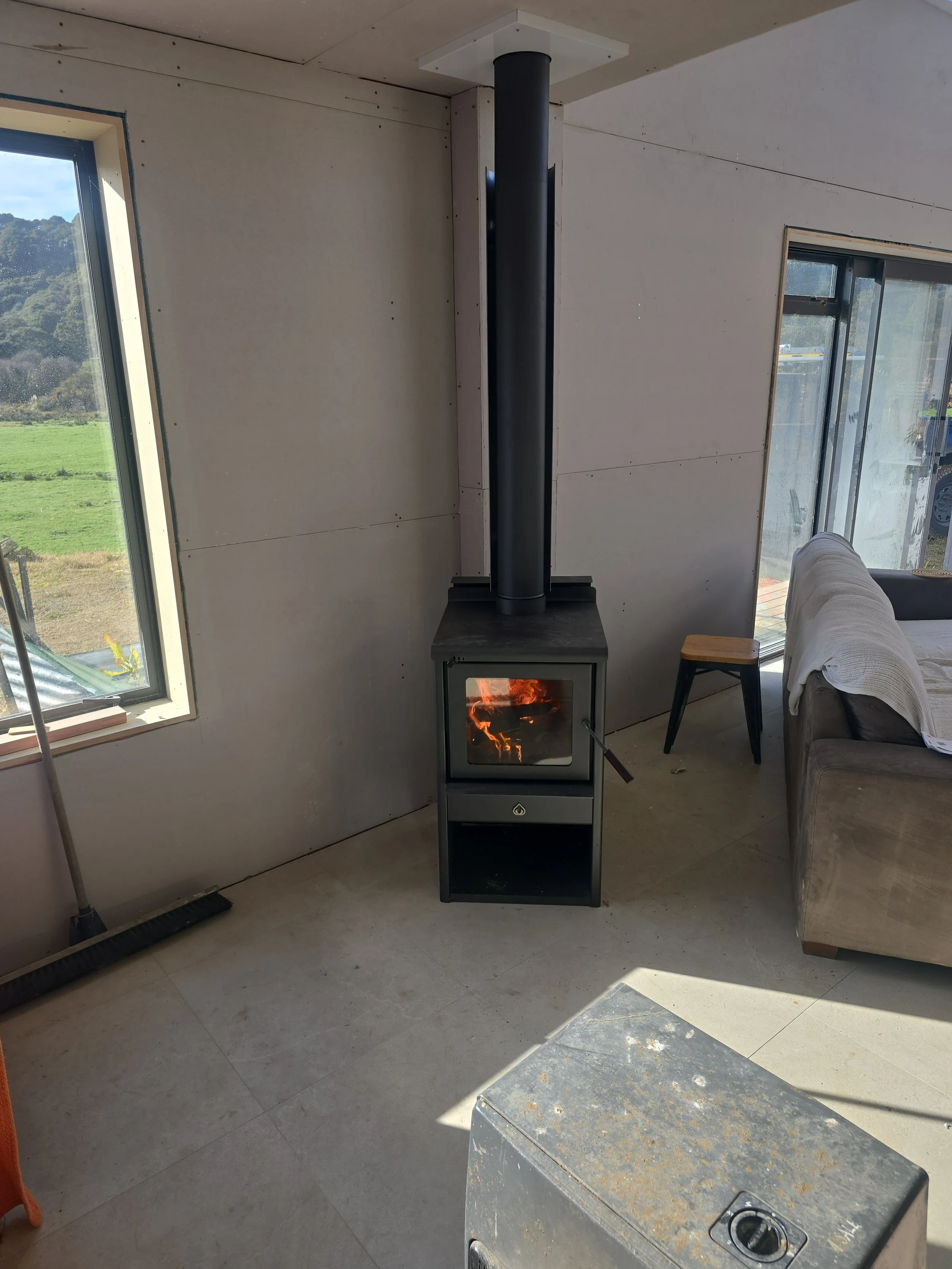 A modern black wood stove with a visible fire inside, situated in a room with unfinished walls, a large window showing greenery outside, and a couch with a white cover on the right side.