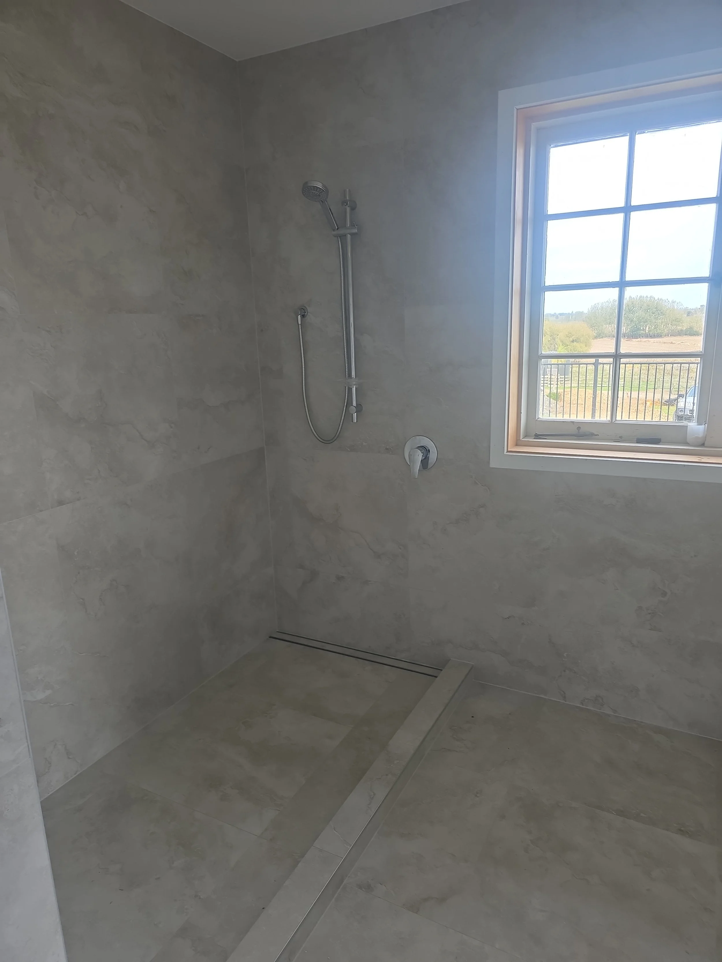 Empty walk-in shower with beige marble tiles, a handheld showerhead, and a window showing an outdoor landscape.