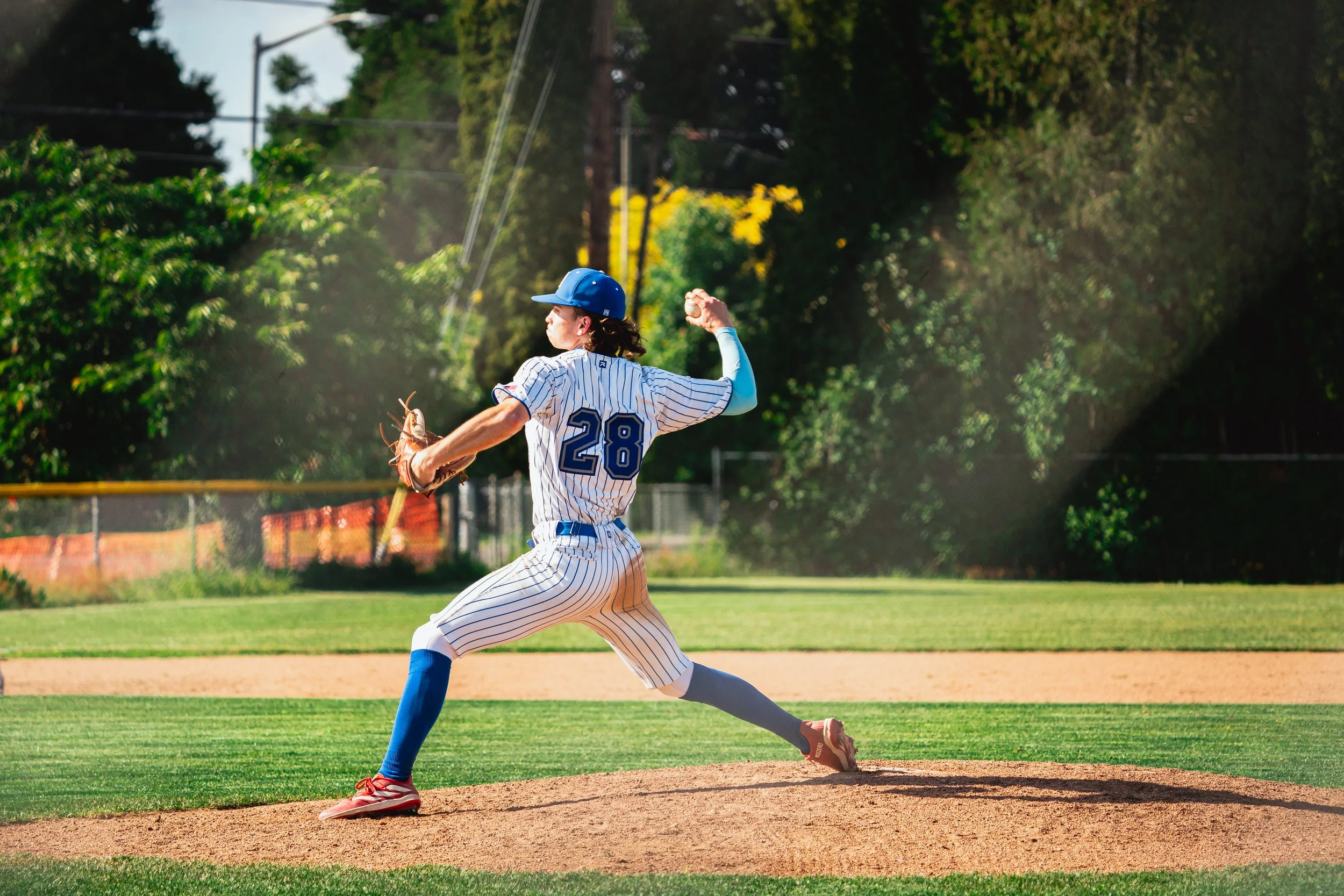 A baseball player in a pinstriped uniform with the number 28 on the back is pitching on a baseball field during daytime. He is wearing a blue cap, blue socks, and red cleats, with trees and a fence in the background.