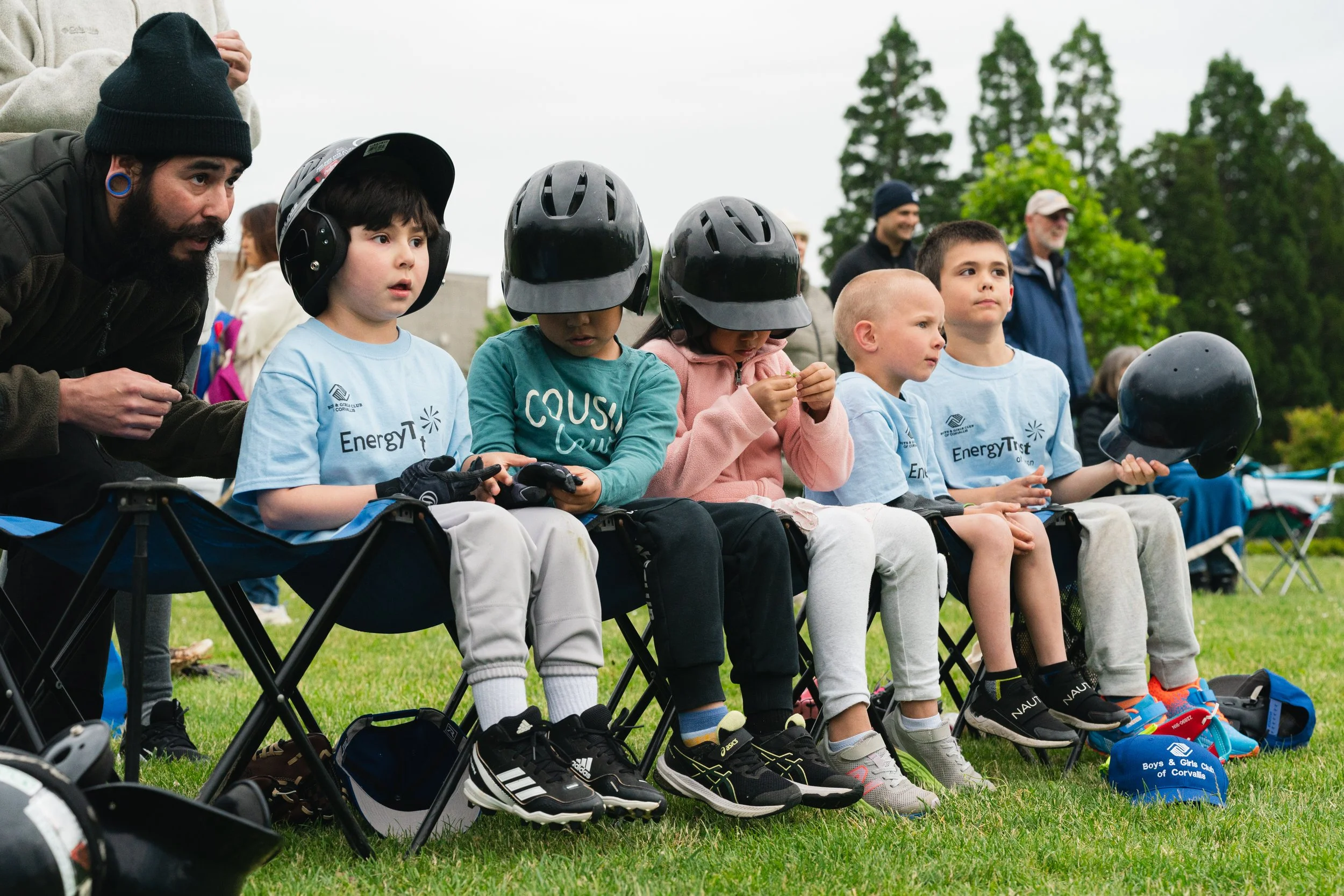 Six children sitting on a bench outdoors, some wearing helmets, with an adult coaching them, and a person in the background. It appears to be a youth event or sports activity.