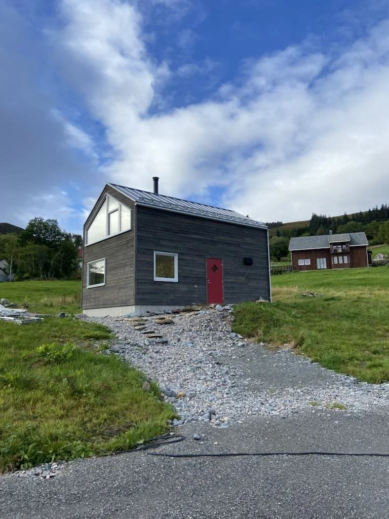 A modern black house with a red door and metallic roof on a grassy hillside under a partly cloudy sky.