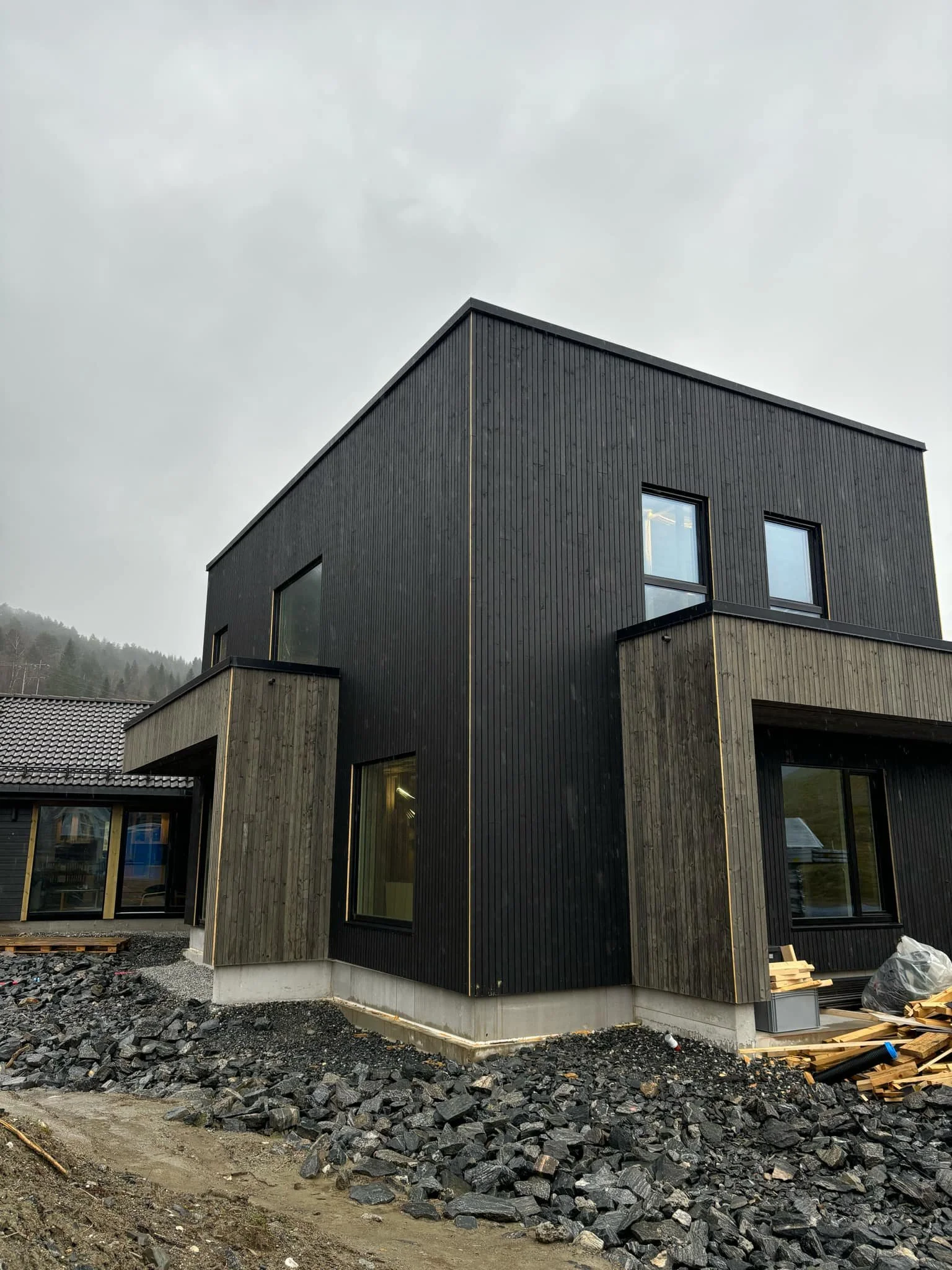 A modern, black and gray house under construction with rocks and dirt in the yard, on an overcast day.
