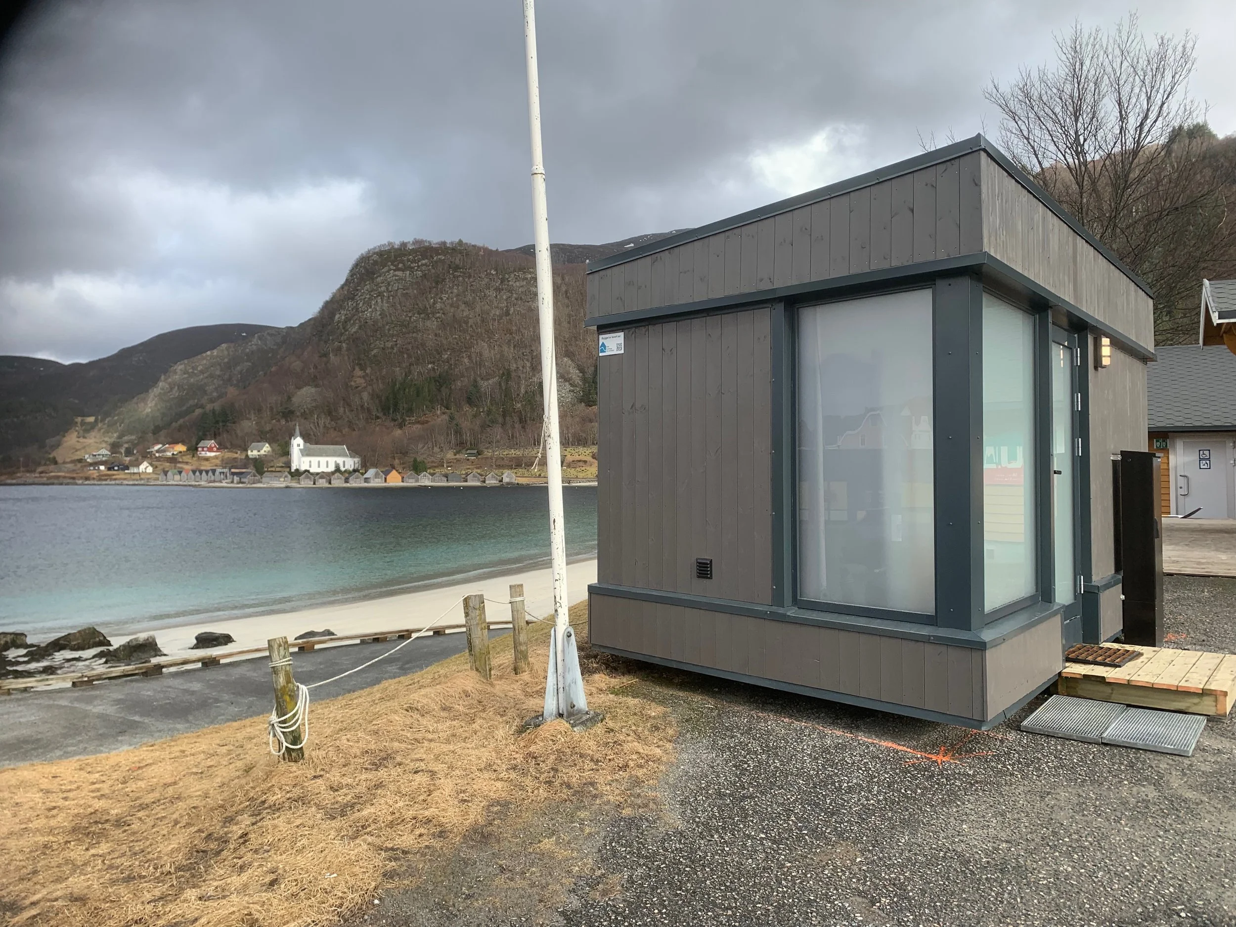 Construction site with a small building made of wood and glass windows, surrounded by construction materials, with snow on the ground and a mountain in the background.