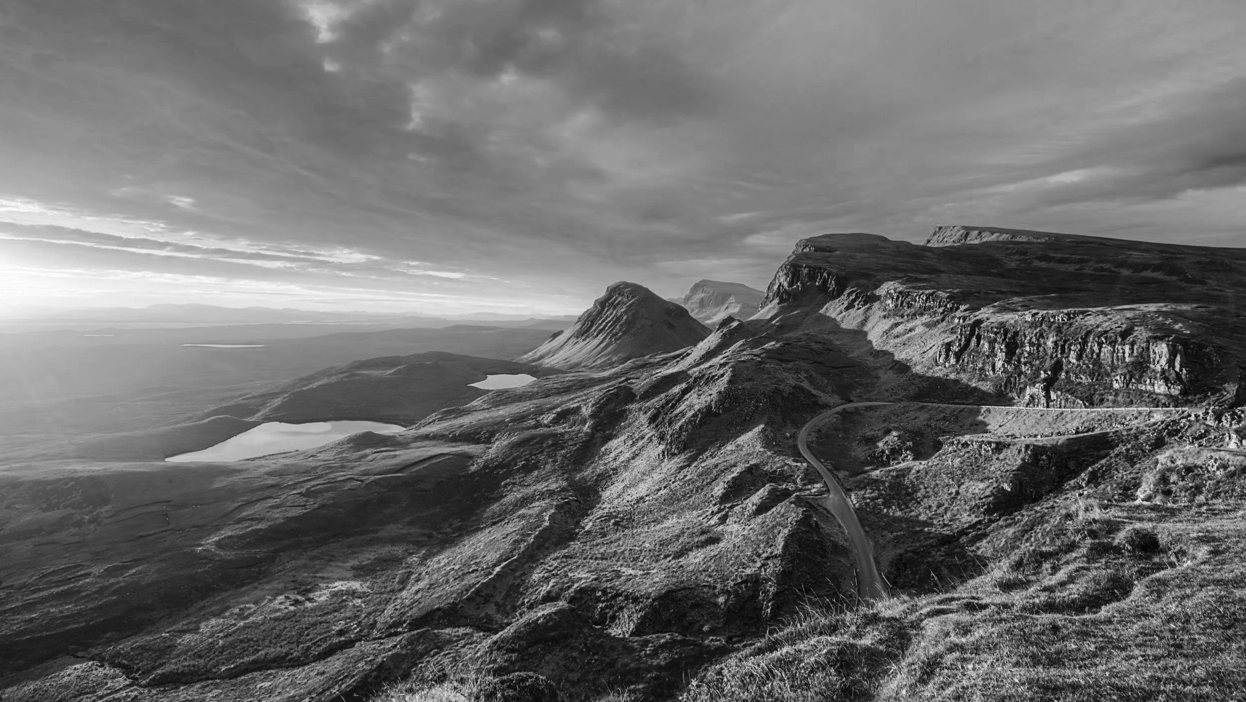 Black and white photo of a mountainous landscape with cliffs, rolling hills, a winding road, and small lakes, under a cloudy sky.