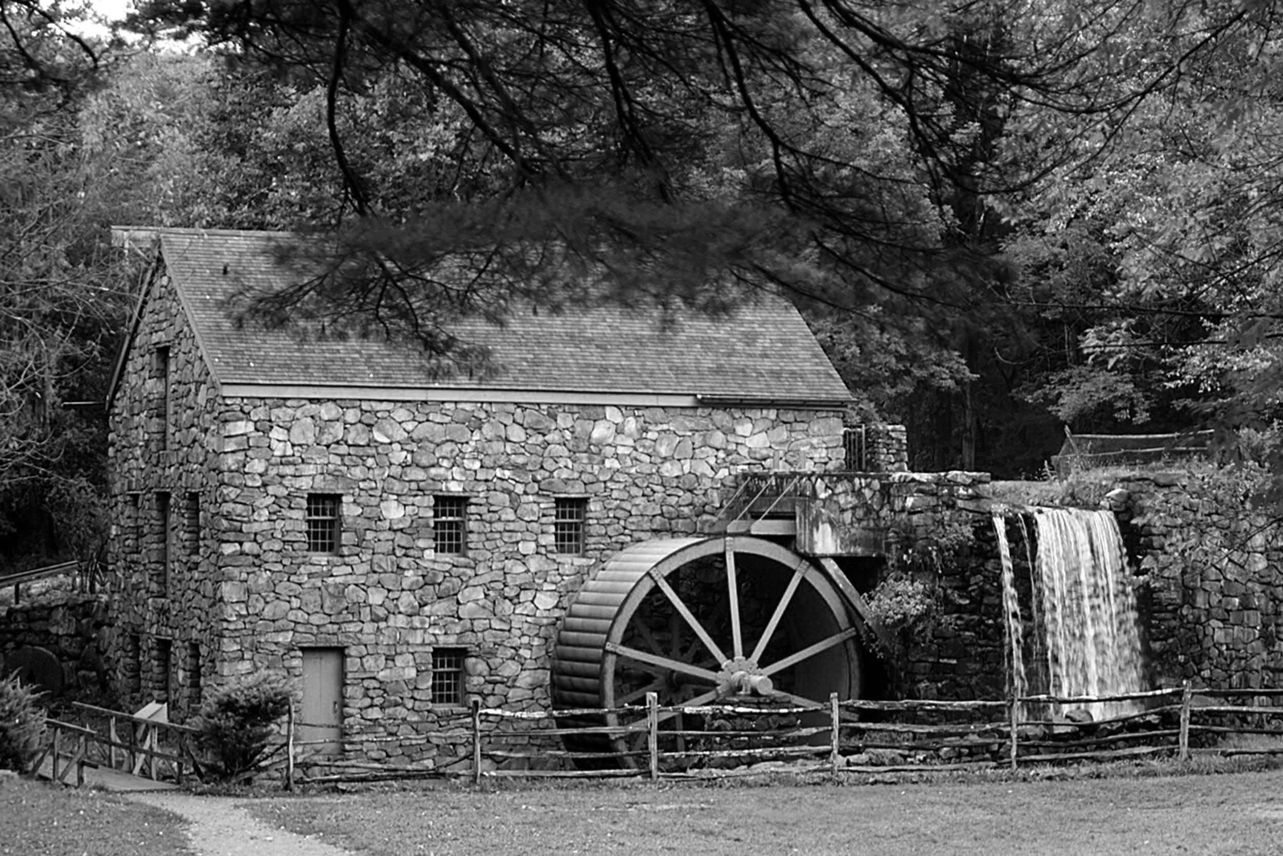 Black and white photo of a stone watermill with a large wooden wheel, a waterfall, surrounded by trees.