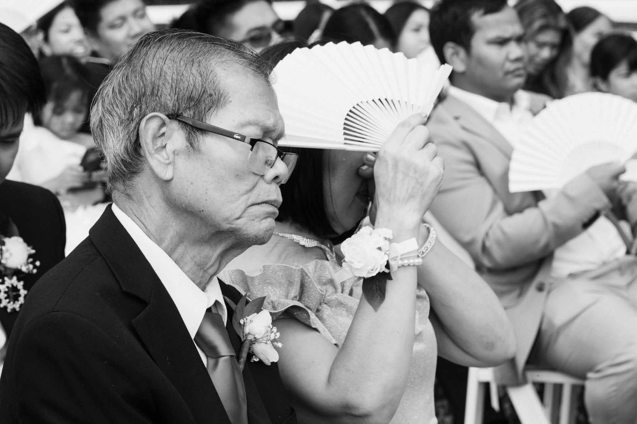 Emotional candid of the groom’s parents during the ceremony, as the mother hides her tears with a fan and the father watches proudly