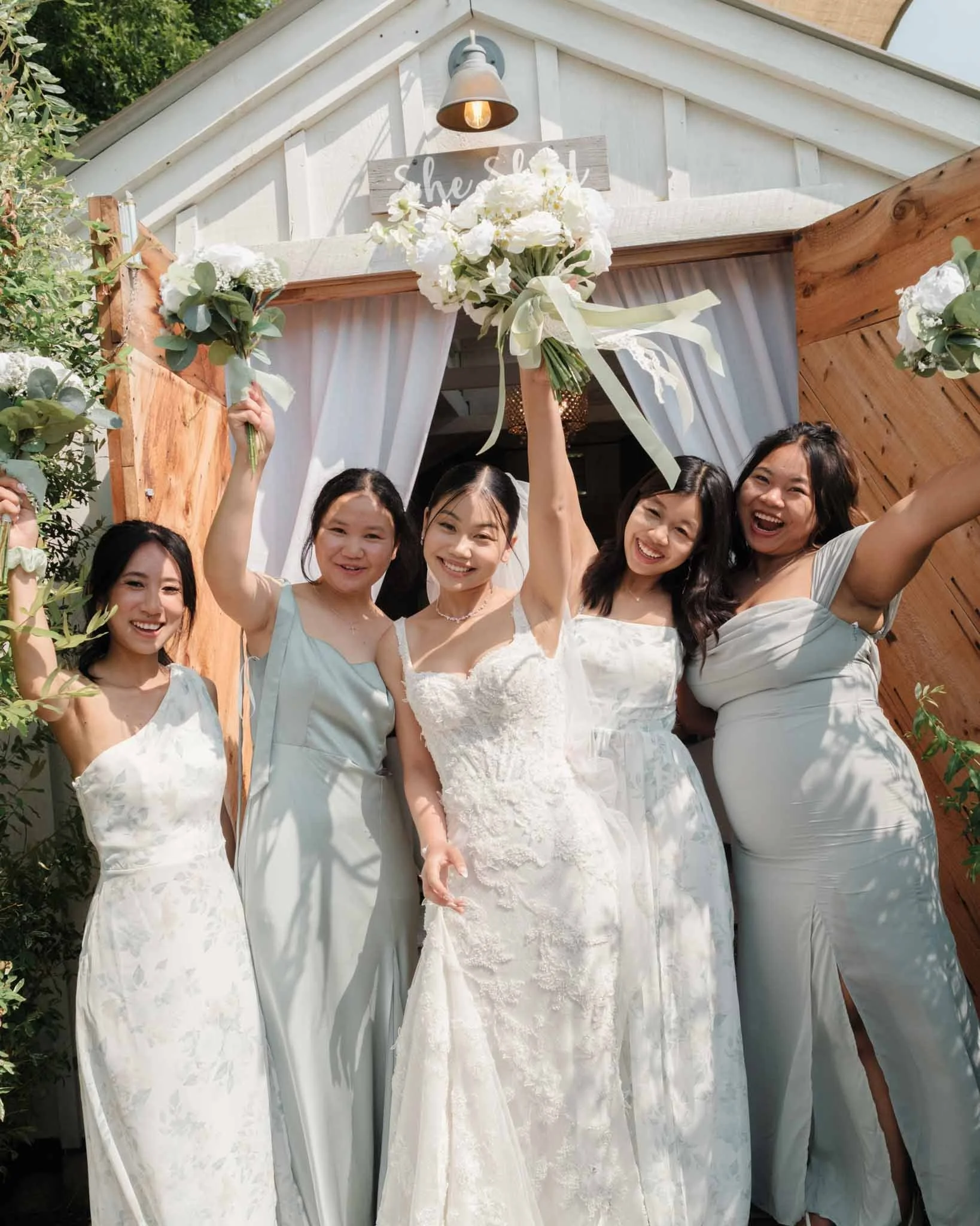 Candid moment of the bride with her bridesmaids in front of their getting-ready shed, cheering with bouquets raised in celebration of the bride