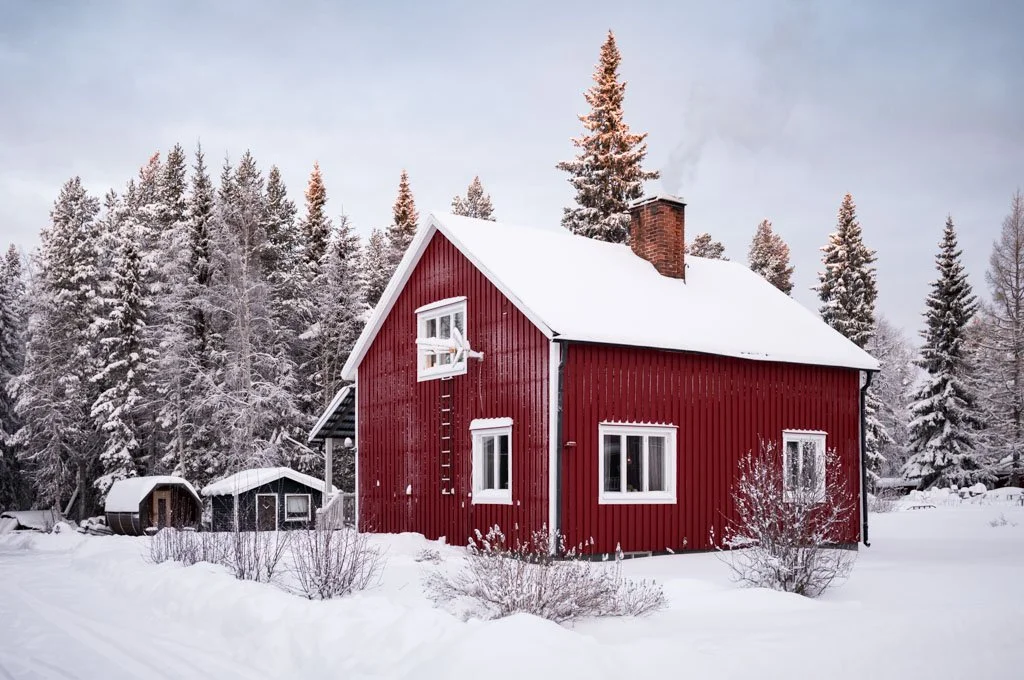 Ein rotes Holzhaus im Winter, umgeben von schneebedeckten Bäumen und Schnee, mit einem Schornstein auf dem Dach.