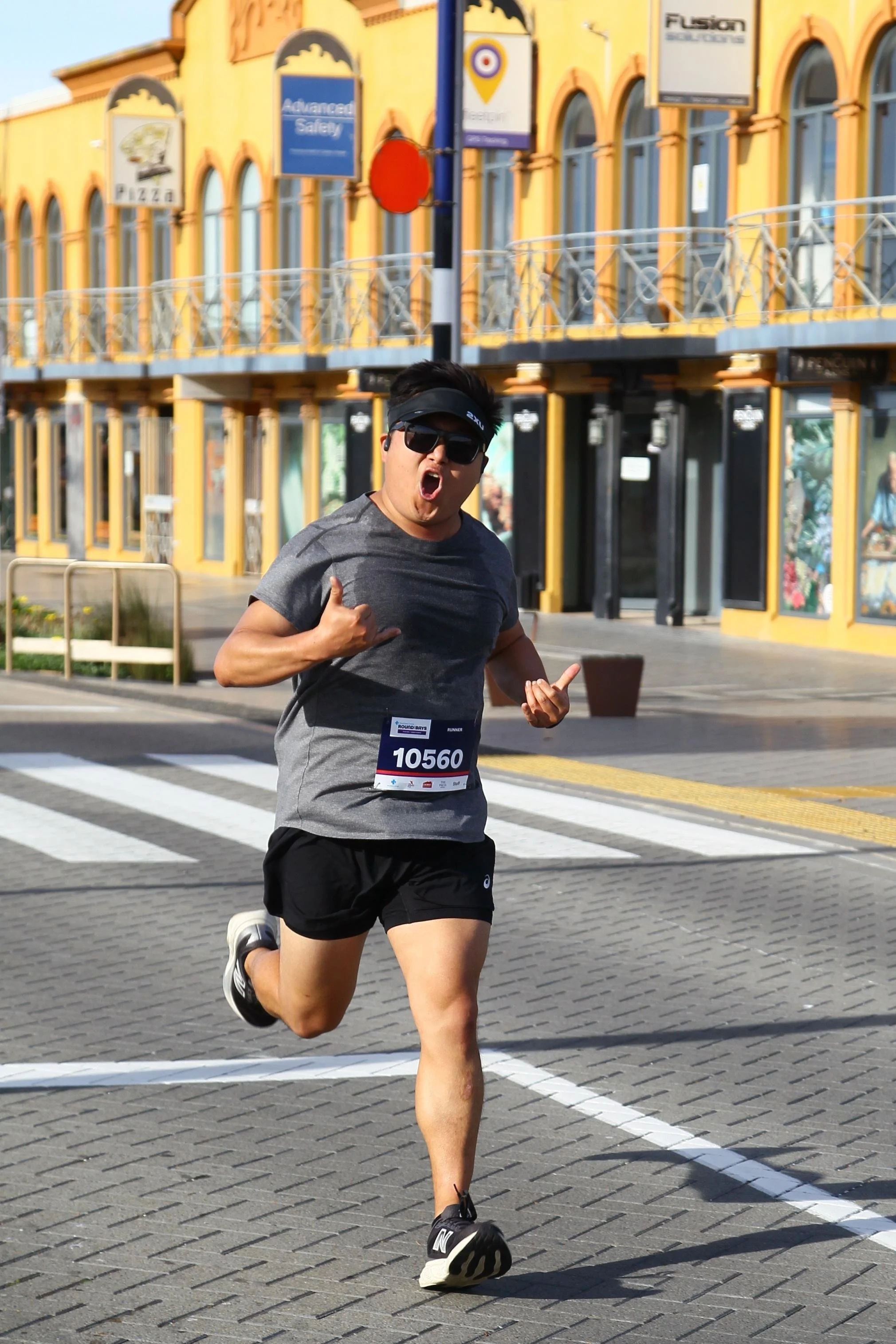 A male runner wearing sunglasses and a gray T-shirt with race bib number 10560 participating in a marathon on a city street with colorful buildings and storefronts in the background.