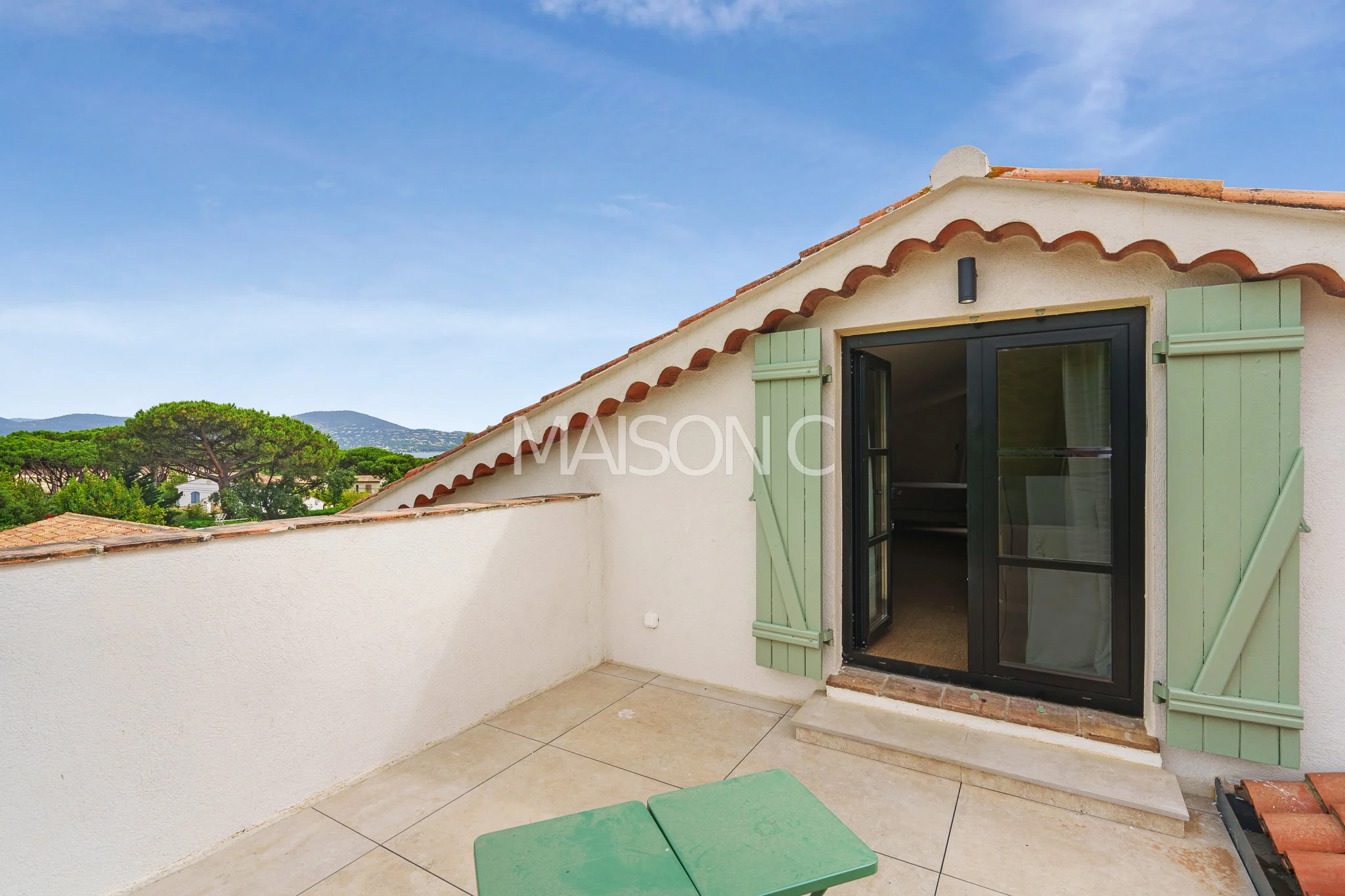 Balcon avec portes-fenêtres noires et volets verts, vue sur des arbres et des montagnes, ciel bleu avec quelques nuages.