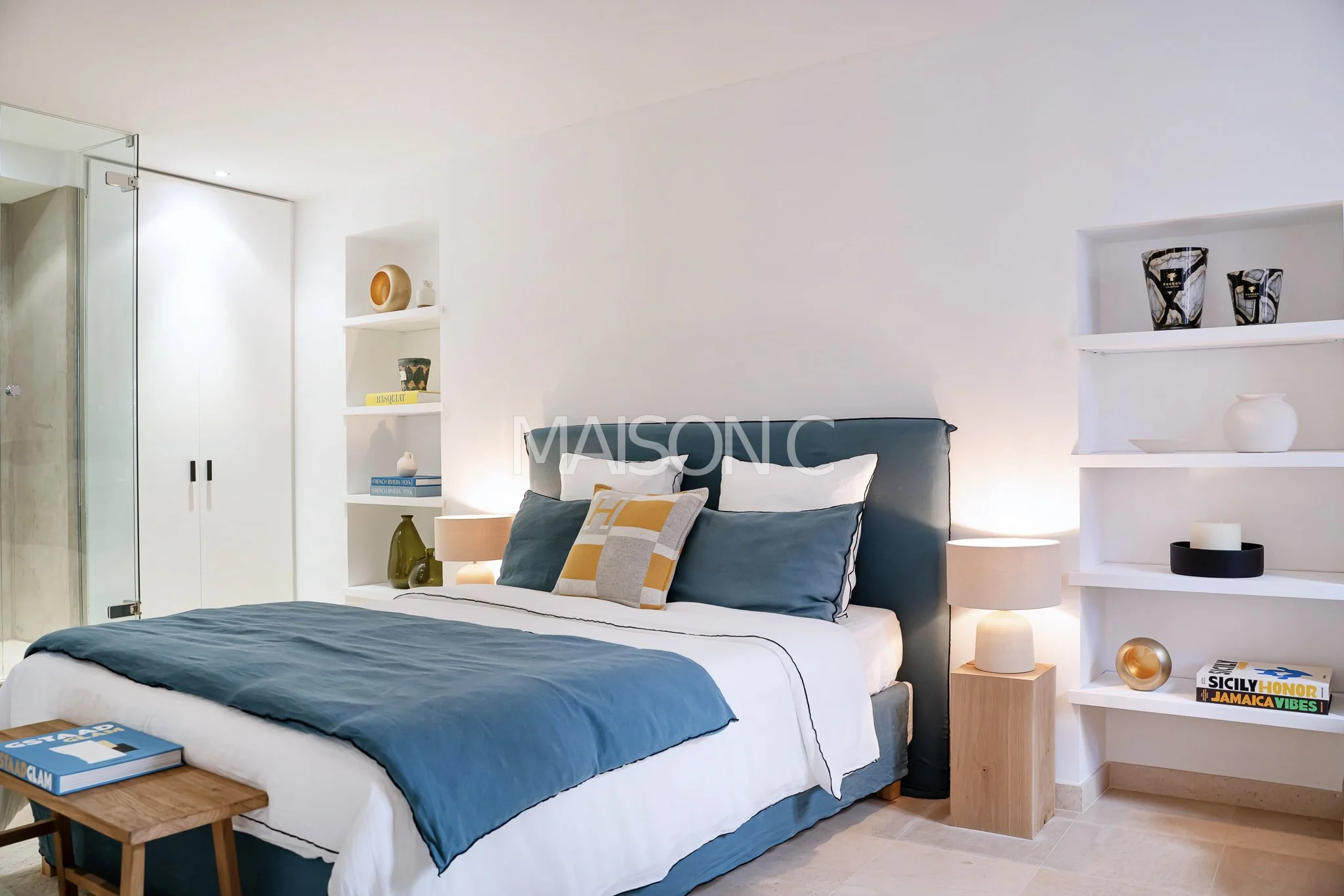 Chambre moderne avec lit à tête rembourrée bleue, oreillers blancs et décoratifs, lampes de chevêt en bois, étagères blanches avec objets décoratifs, et sol en carrelage beige.