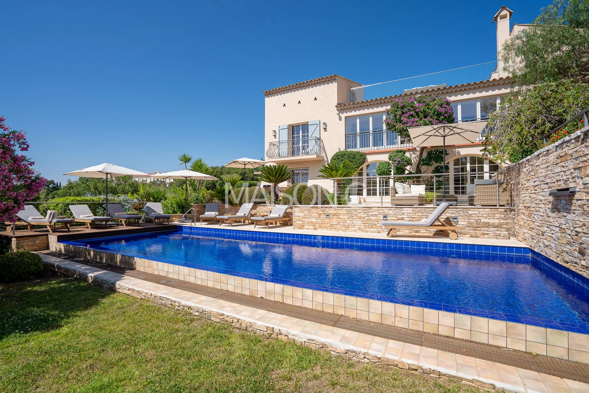 Piscine de style résidentiel avec chaise-longues et parasols sous un ciel clair, entourée de végétation et d'une maison en pierre avec des balcons et un jardin