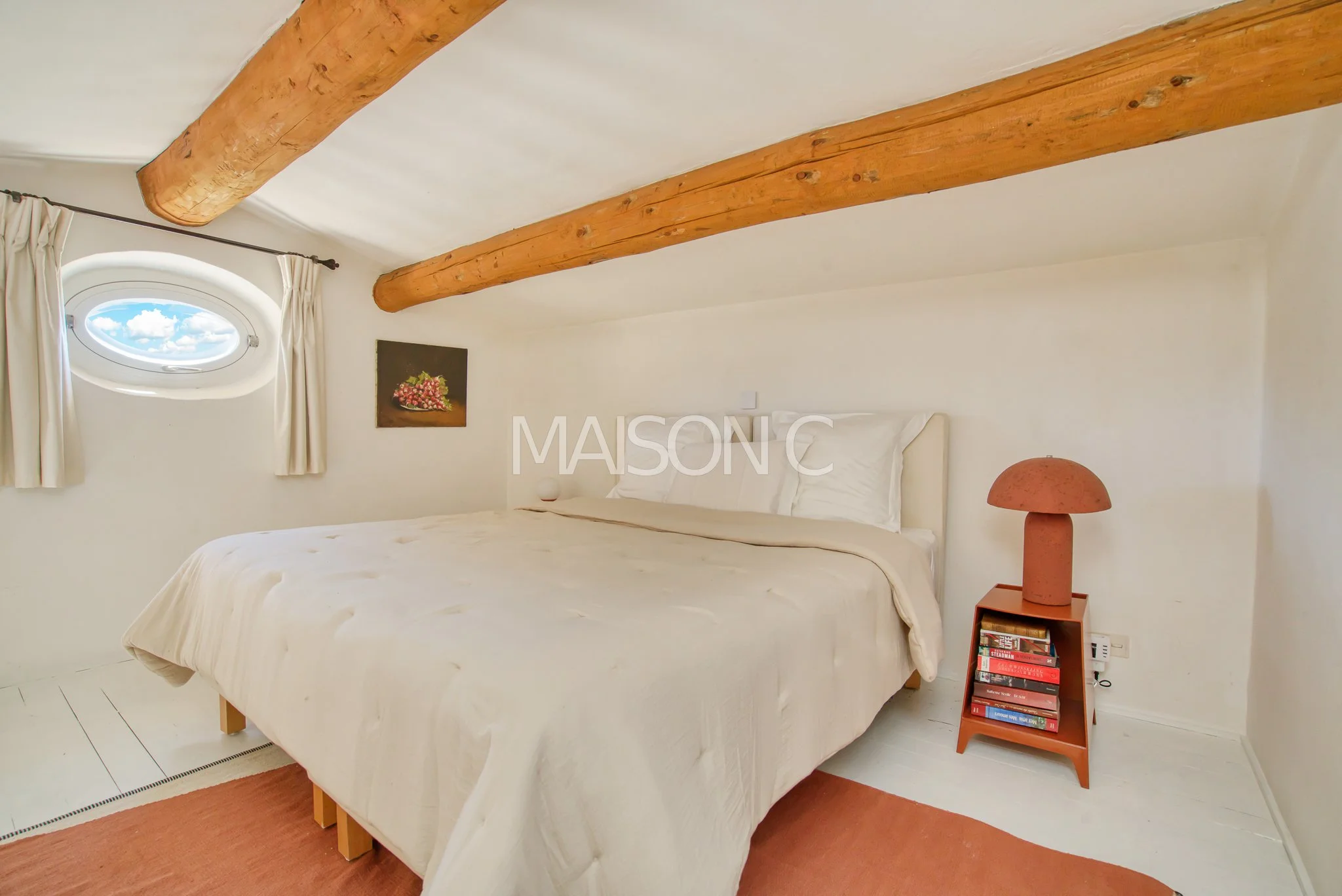 Chambre à coucher avec lit blanc, lampadaire en forme de champignon, petite étagère avec livres, fenêtre ronde avec vue sur le ciel, poutres en bois au plafond, murs blancs.