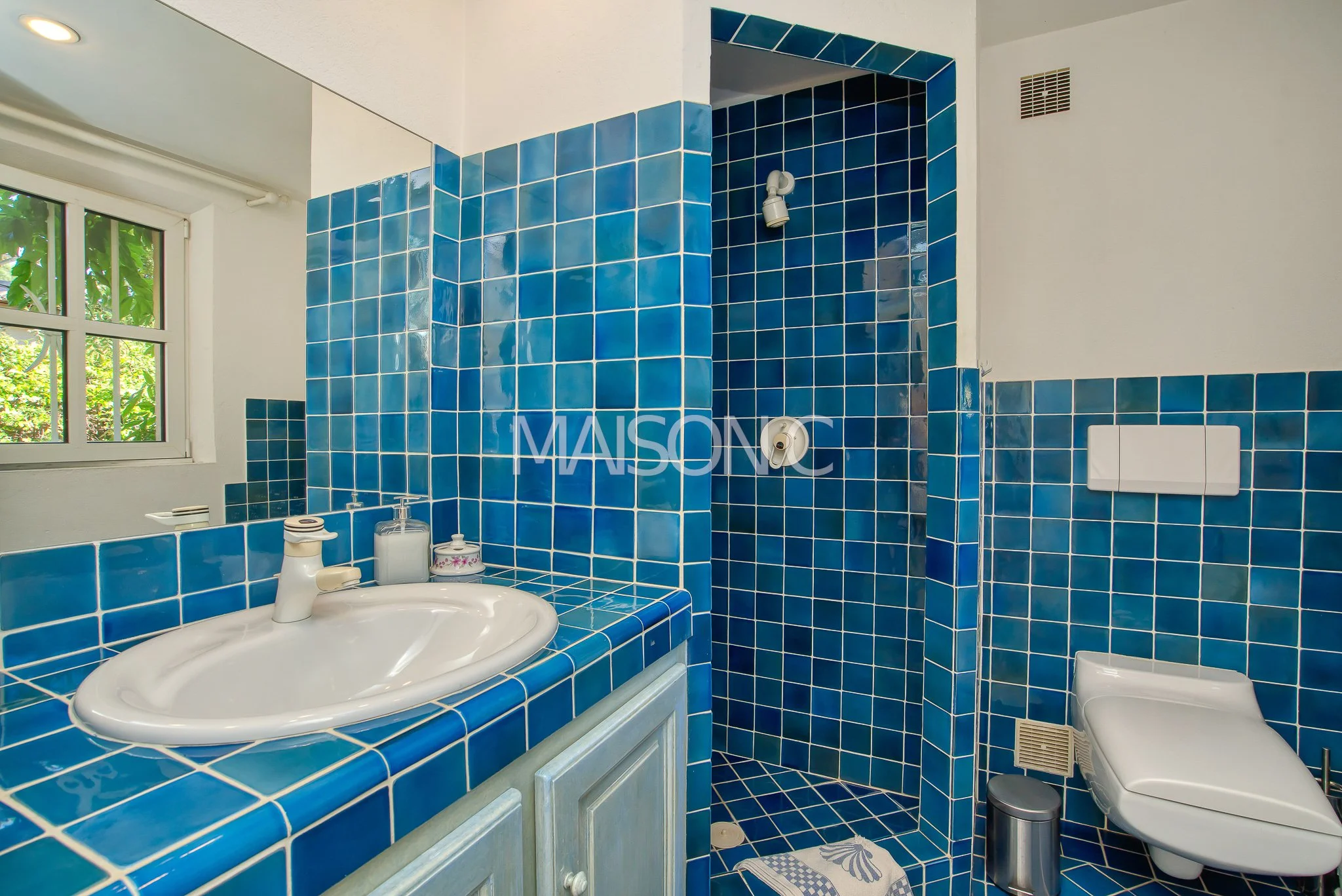 Salle de bain avec murs et lavabo en carreaux bleus, avec une fenêtre laissant entrer la lumière naturelle, comprenant un lavabo blanc avec un miroir au-dessus, une douche en coin, et un WC suspendu.