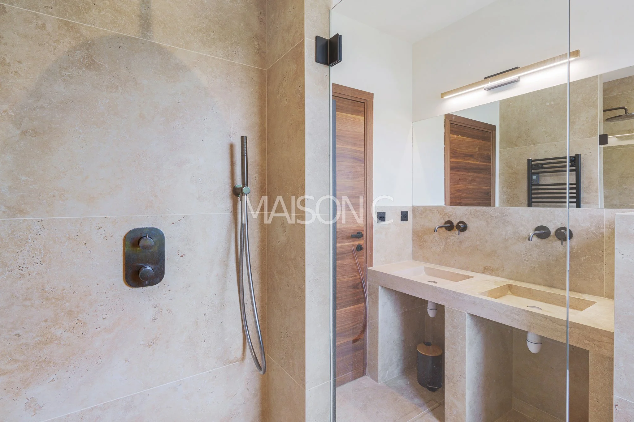 Salle de bain moderne avec un mur en pierre beige, miroir, lavabo double en pierre, porte en bois, ventilos, et radiateur noir.