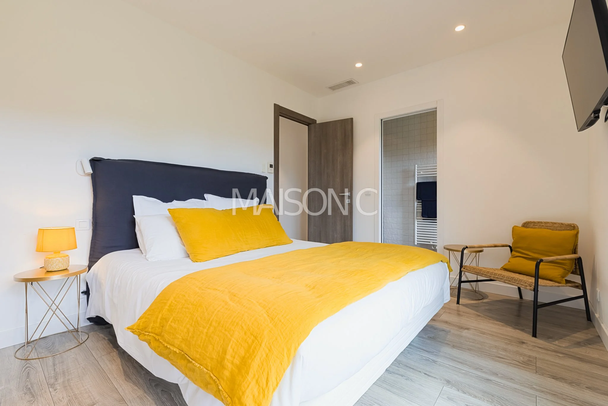 Chambre moderne avec un lit à draps blancs, couverture et oreillers jaunes, petite table de chevet avec lampe orange, fauteuil en bois avec coussin jaune, porte entrouverte vers une salle de bain, télévision murale.