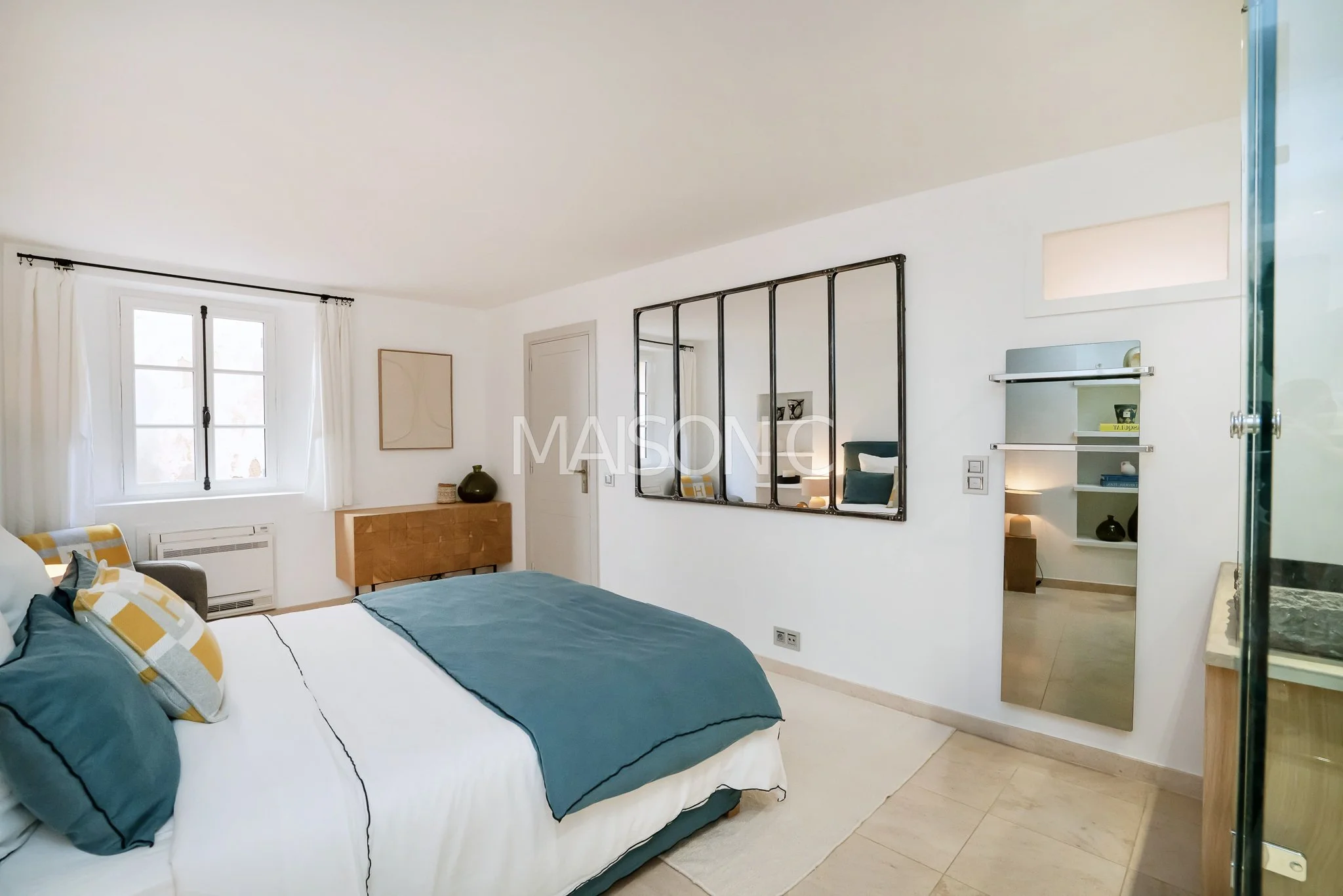 Chambre avec lit double, oreillers colorés, fenêtres avec rideaux blancs, miroir mural, mobilier en bois, décoration neutre.