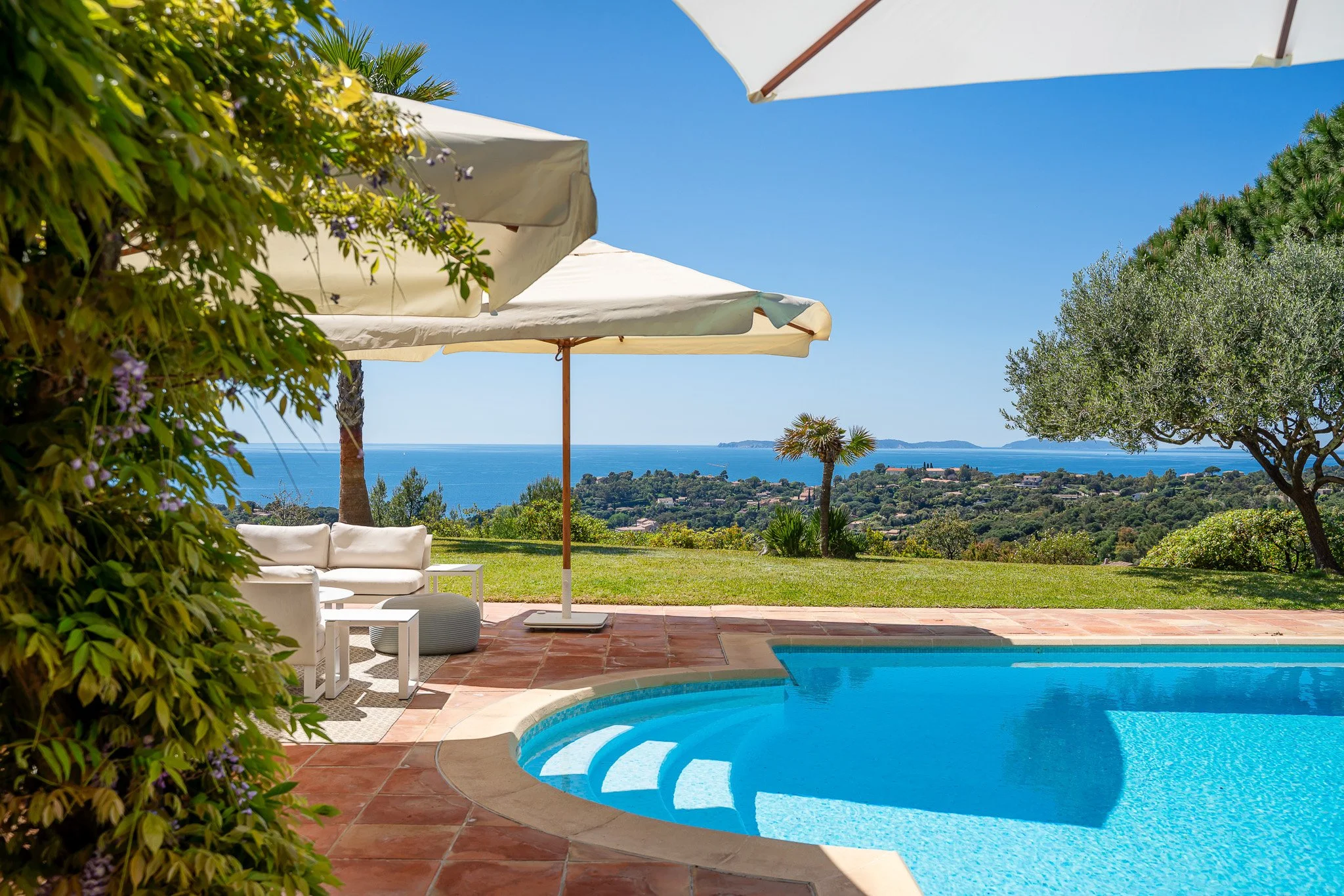 Piscine avec terrasse en terre cuite, parasols blancs, mobilier de jardin blanc, vue sur la mer, arbres et végétation tropicale dans un paysage ensoleillé.