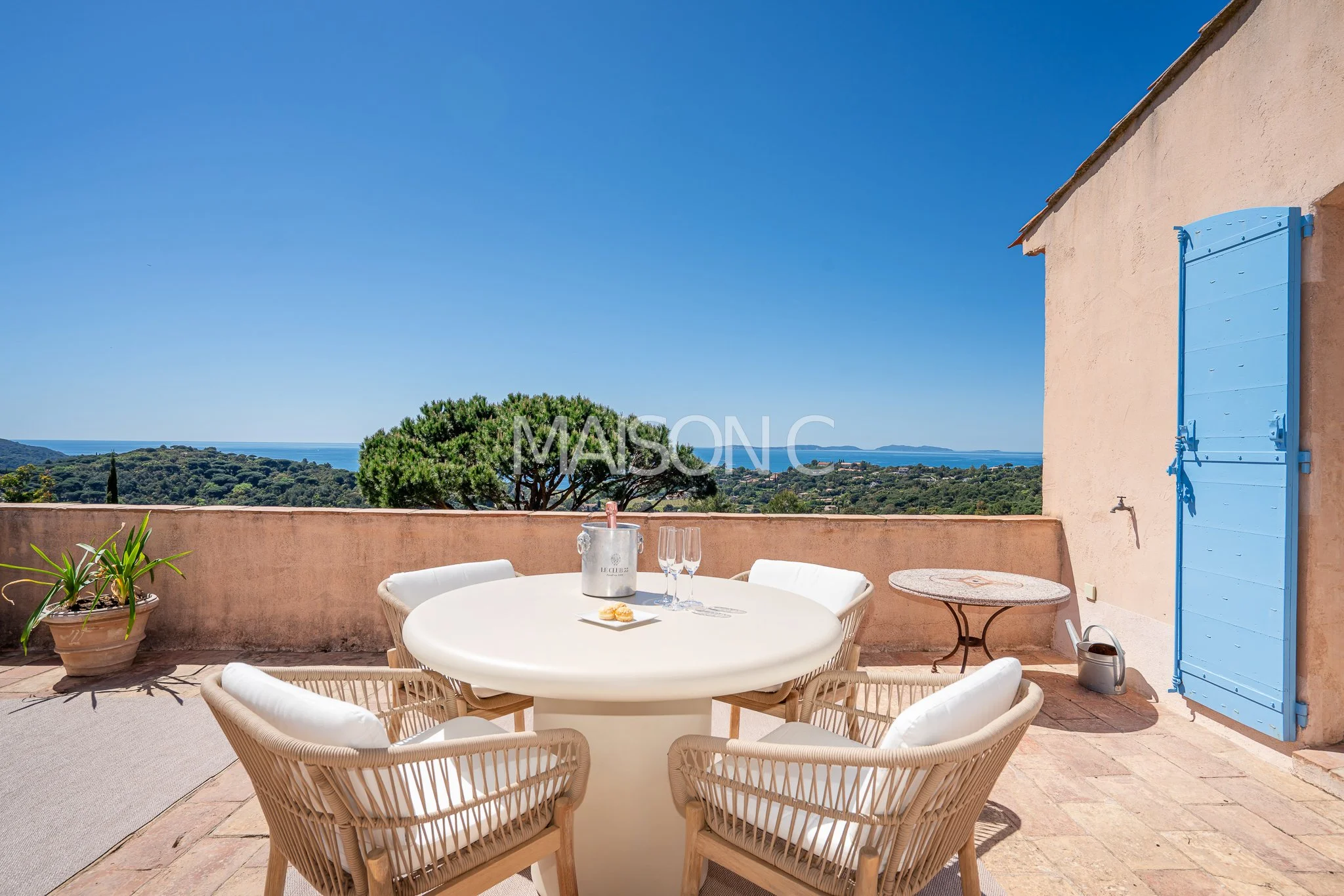 Terrasse ensoleillée avec vue sur la mer, table ronde blanche avec chaises, une bouteille de champagne, deux flûtes, et un petit plat, côté maison avec un volet bleu