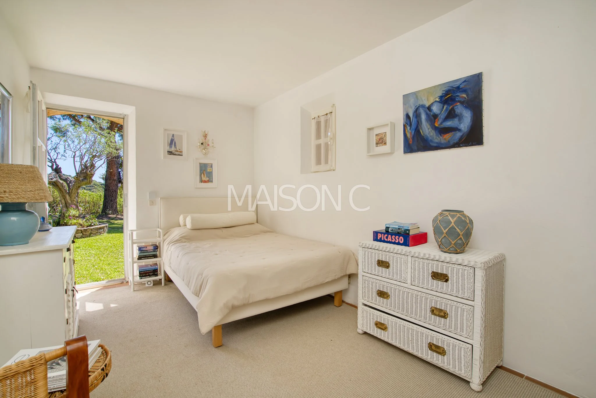 Chambre lumineuse avec lit blanc, dresser blanc, décoration artistique sur mur, porte ouverte sur jardin avec arbres et pelouse.