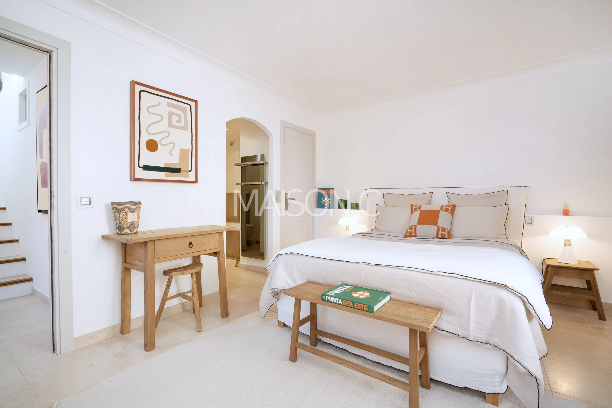 Chambre à coucher blanche avec lit grand format, coussins orange et beige, tables de chevet en bois, lampes, décoration moderne, tableau abstrait, coffre en bois, et petits objets décoratifs.