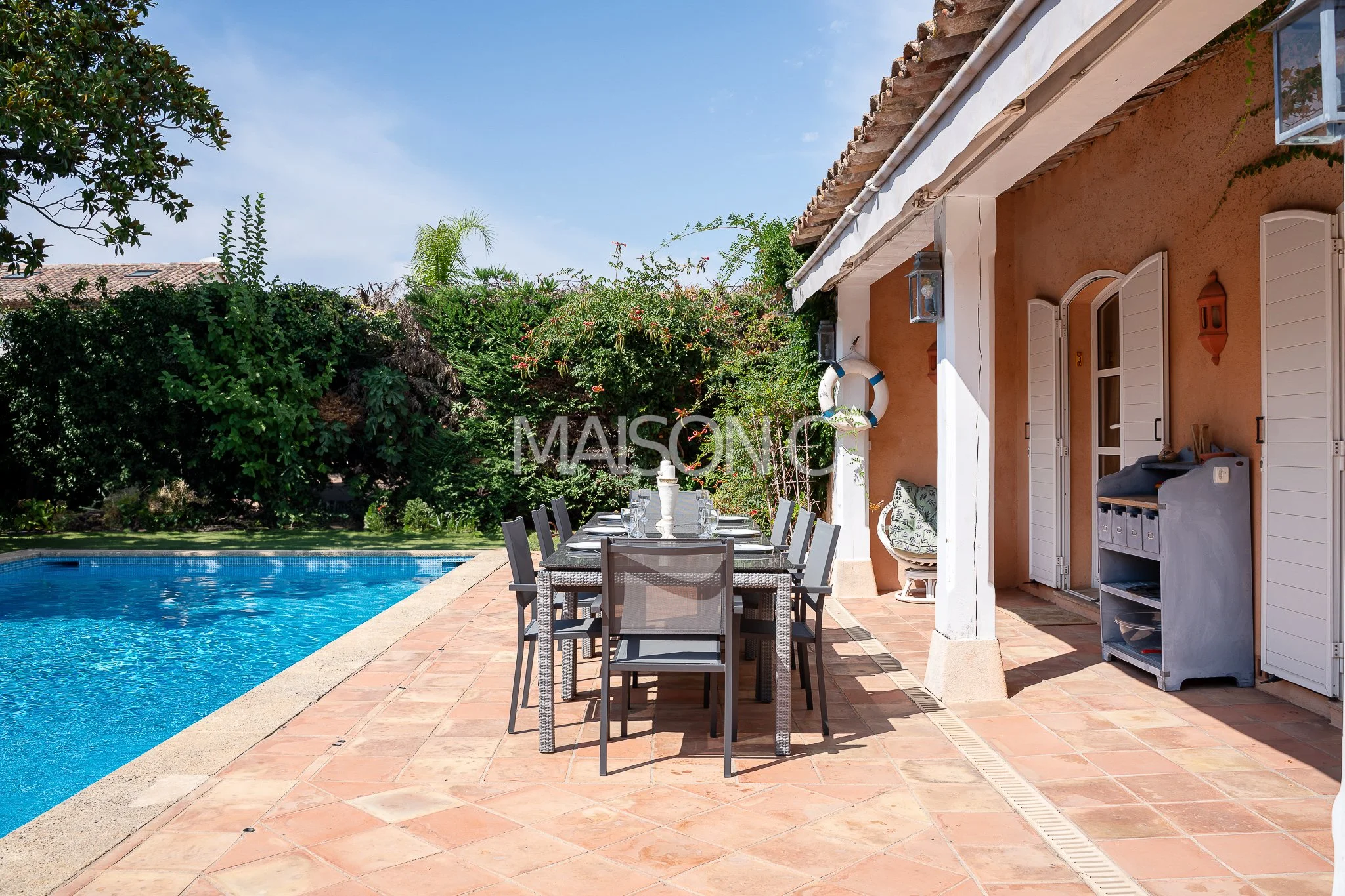Terrasse ensoleillée avec table et chaises près d'une piscine, entourée de végétation luxuriante, maison en zone tropicale.